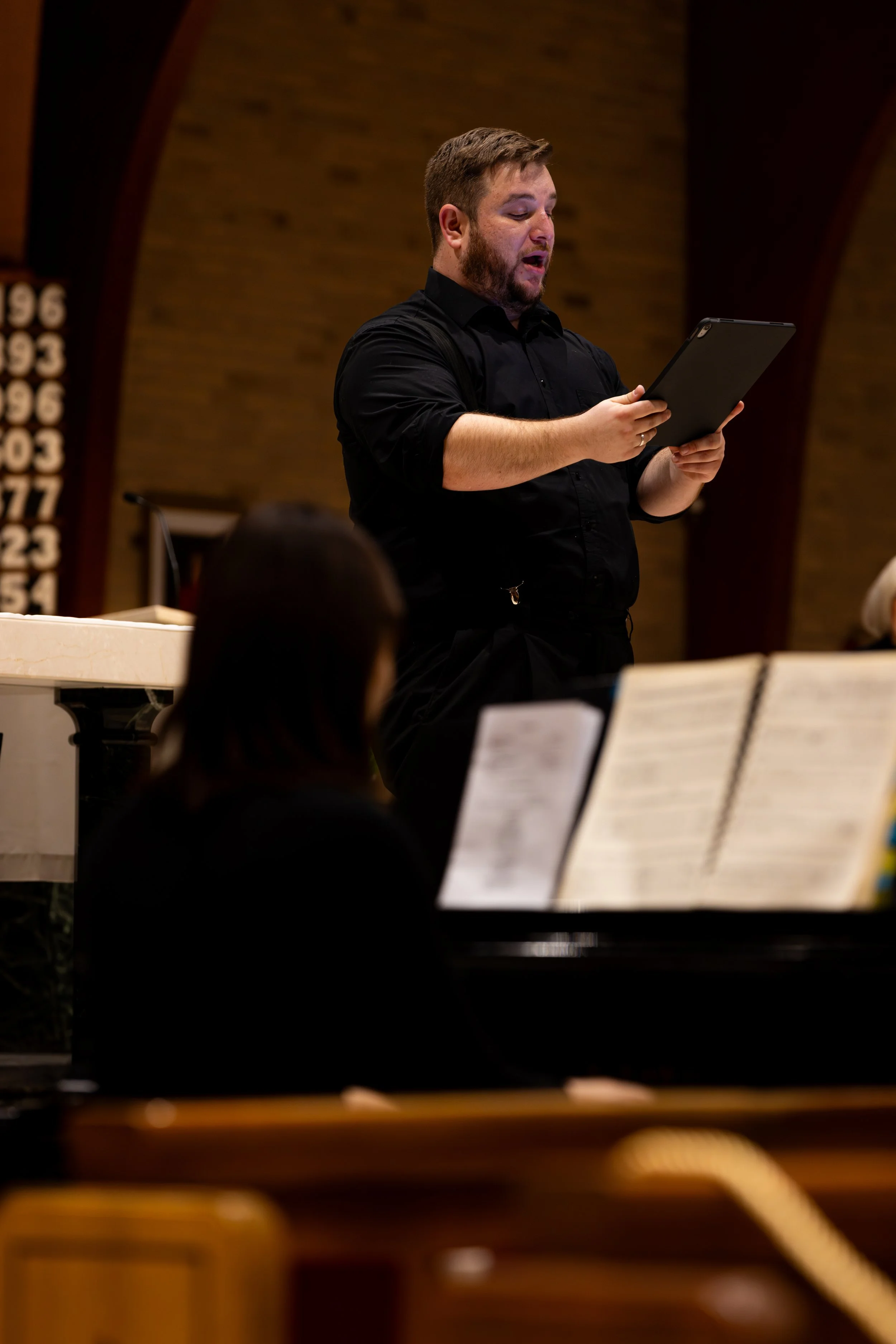 A man with brown hair wearing a black shirt holds music as he sings a solo.