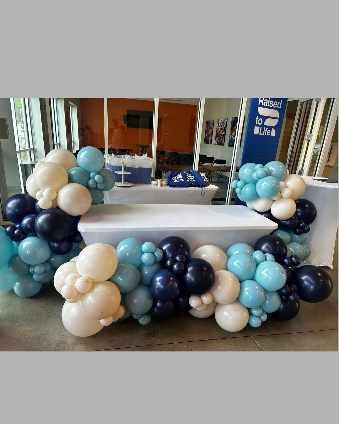 Desk decorated with clusters of white, light blue, dark blue, and cream balloons at a booth for a charity event with a sign that says "Raised to Life."