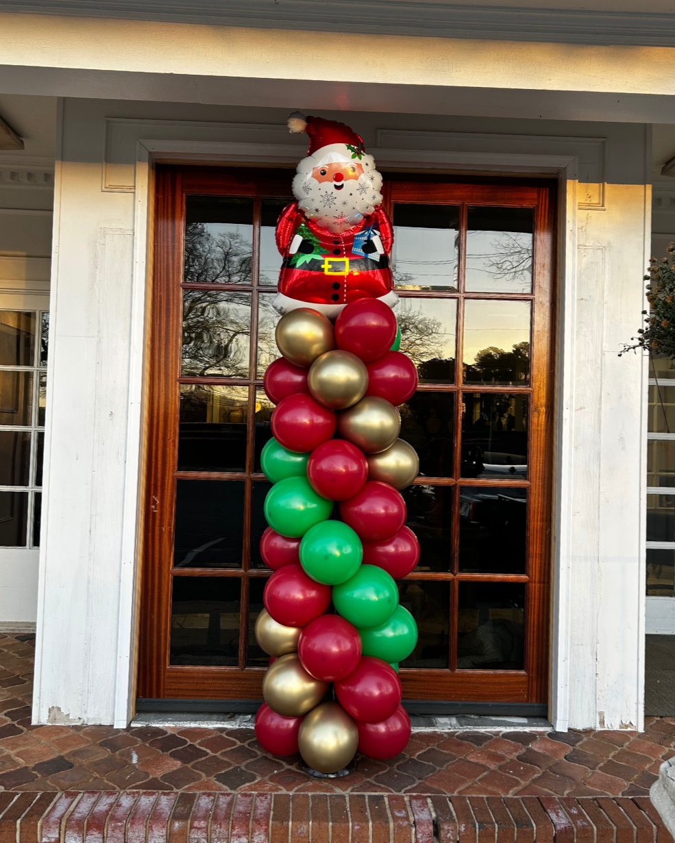 A column of red, green, and gold balloons topped with a cheerful Santa Claus balloon decoration, placed in front of a wooden door with glass panes on a brick porch.