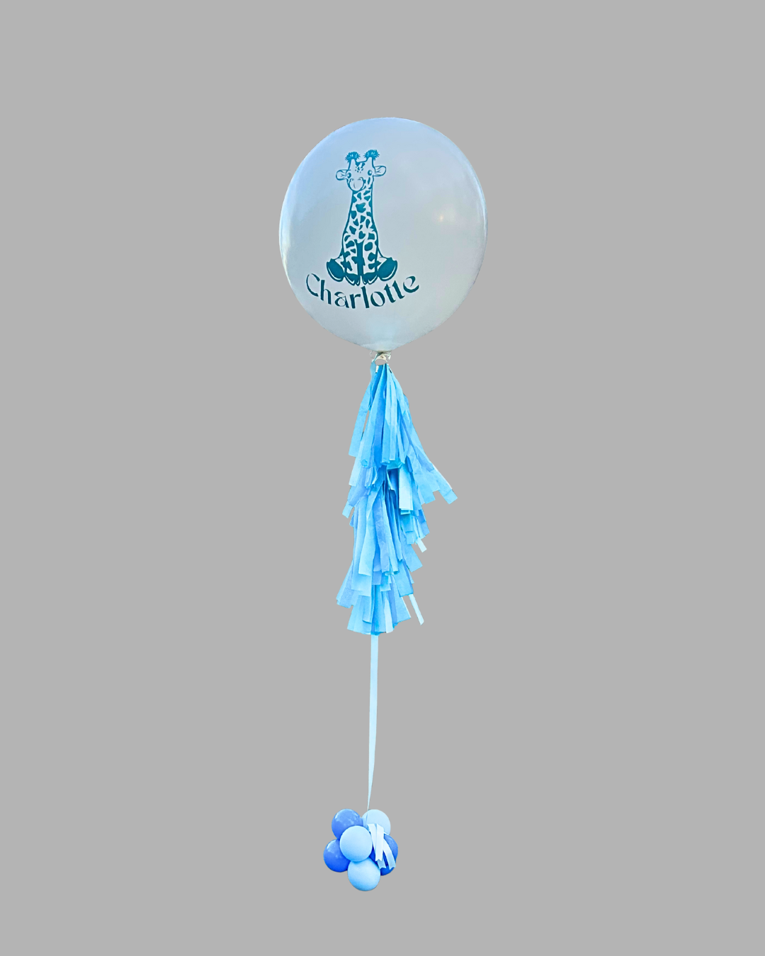 Blue and white balloon with a giraffe illustration and the name 'Charlotte' printed on it, attached to blue paper tassels and additional balloons at the bottom.