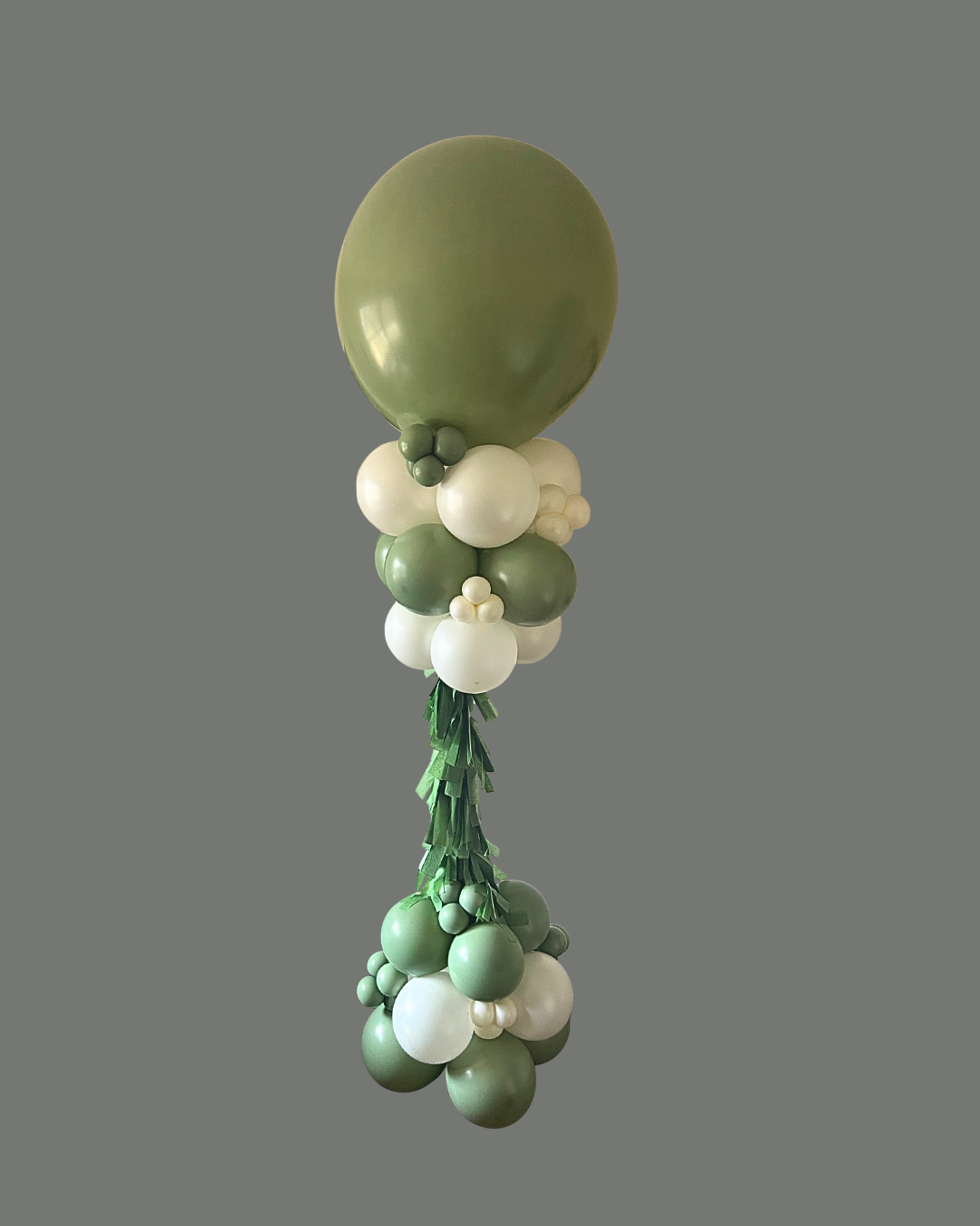 A balloon arrangement featuring a large green balloon at the top, surrounded by smaller white and green balloons, with green paper fringe hanging below, forming a decorative bouquet.