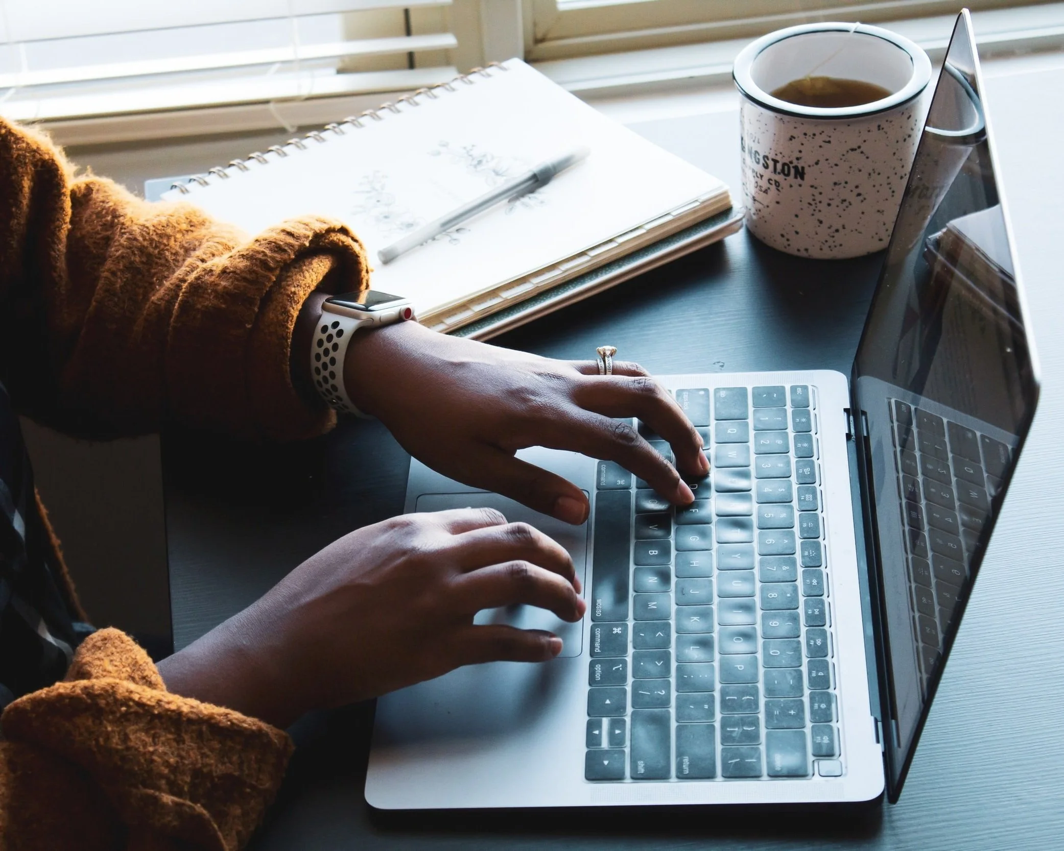 Person using a laptop with a smartwatch on their wrist, a notebook with a pen, a coffee mug, and some papers on a desk by a window.