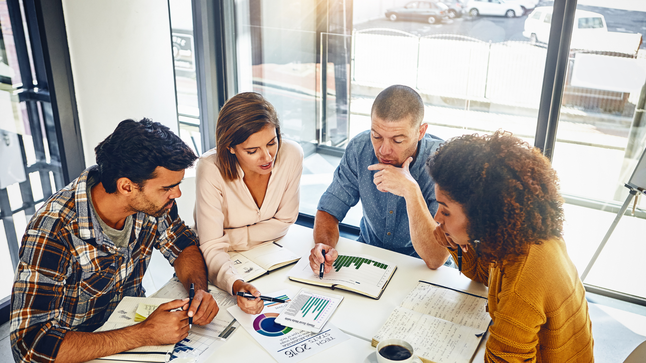 Group of five professionals in a meeting room gathered around a table with charts, notebooks, and documents, discussing work.