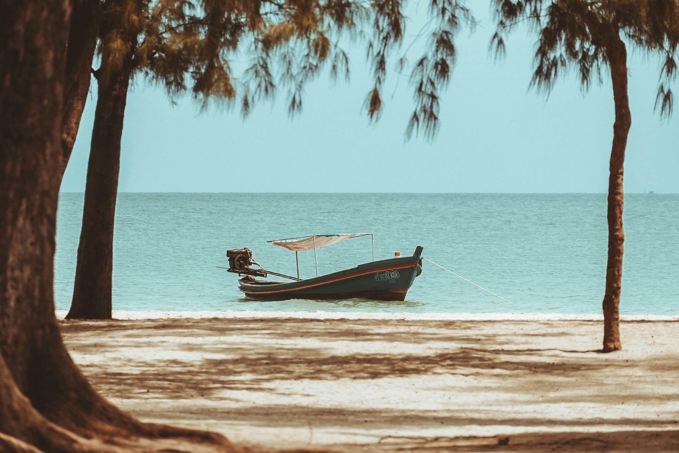 A boat anchored in the water near a beach, framed by trees with shade on the sandy shoreline.