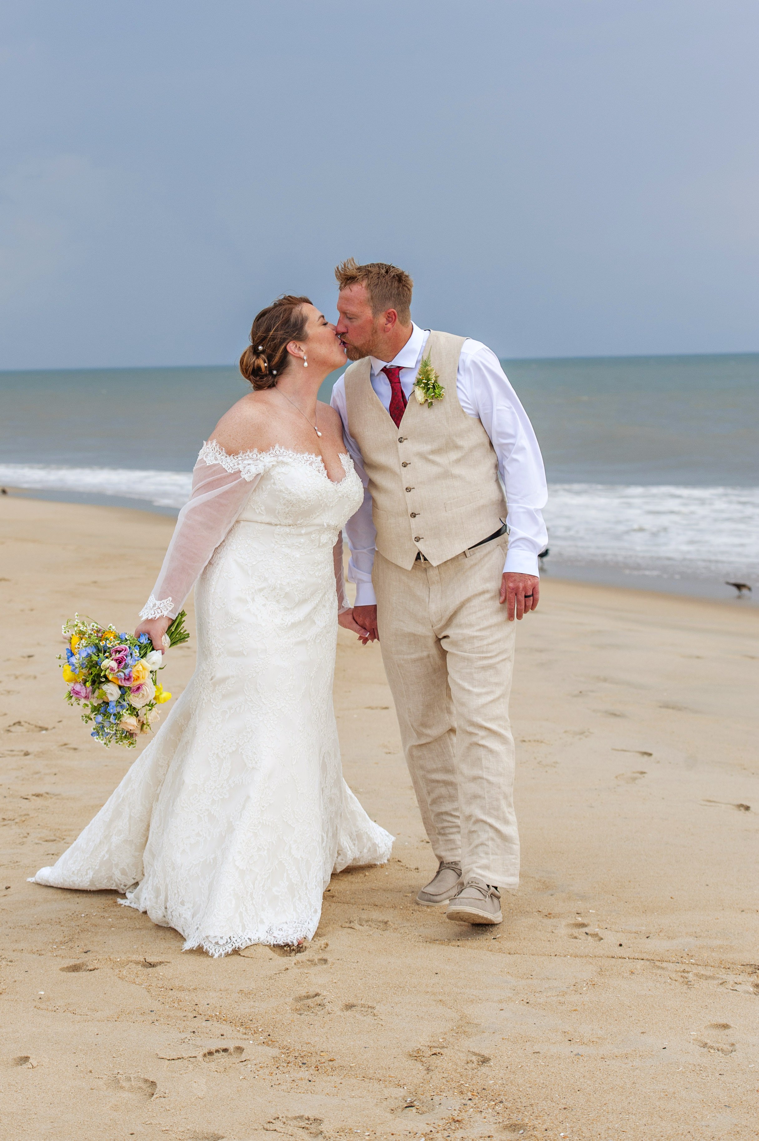 Bride and groom kissing on OBX beach holding colorful bouquet for preservation by Beaux’s Botanical Designs
