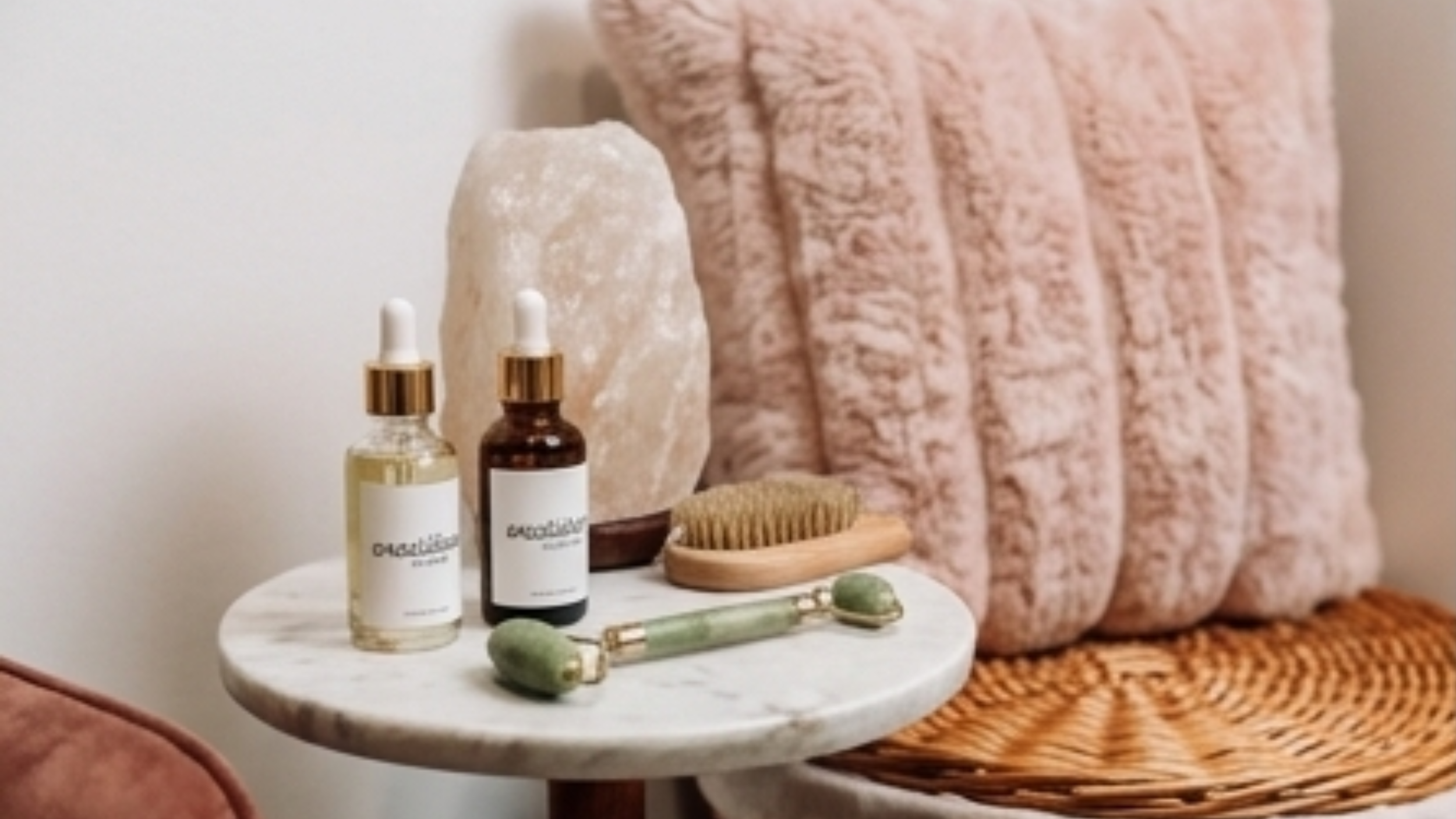 A round marble side table with skincare bottles, a salt lamp, a wooden body brush, and a jade roller, next to a pink textured chair with a woven seat