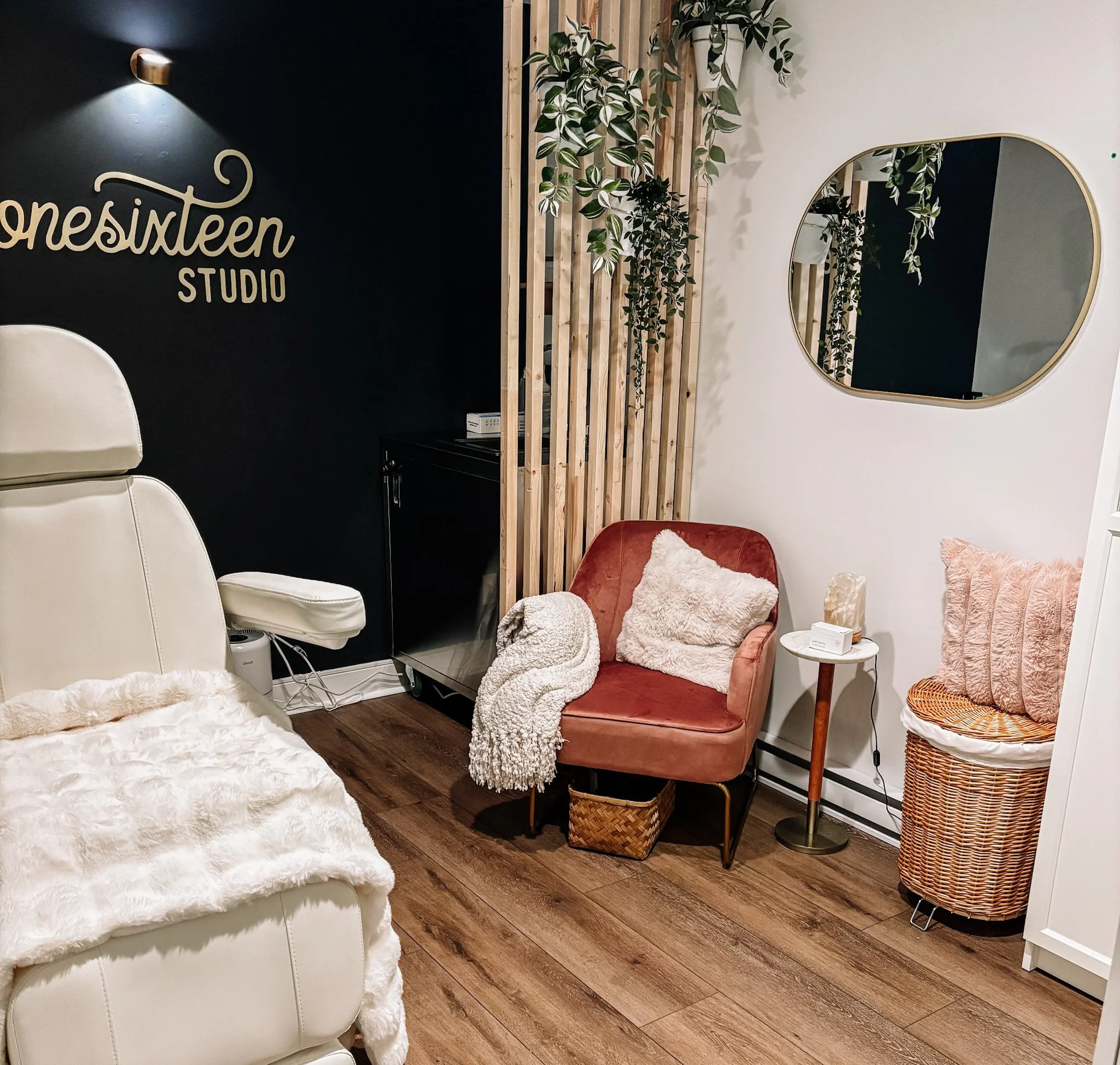 A cozy room with a white reclining chair covered with a soft cream blanket, a pink armchair with a fluffy cream pillow, a small side table with a lamp, and a large wicker laundry basket. There is a black wall with the words 'Onesixteen Studio' and wooden slats with hanging plants, a mirror on the wall reflects part of the room, and the floor is wood.