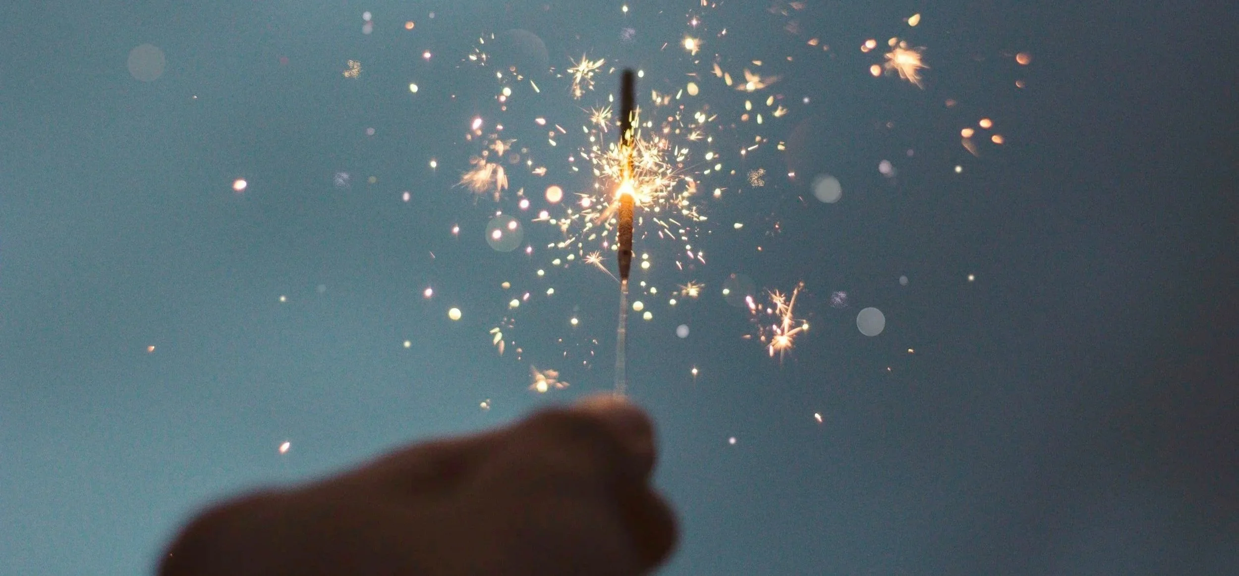 A hand holding a lit sparkler with sparks flying against a blue sky.