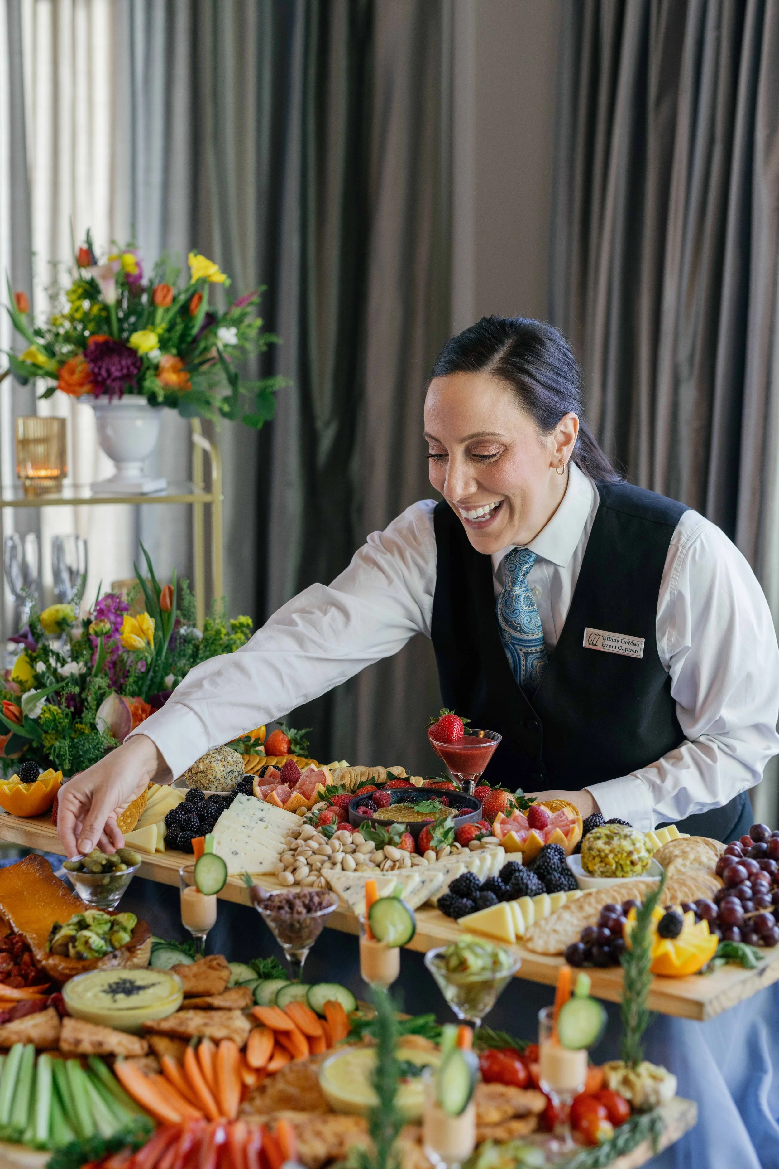 A woman in a uniform smiling as she reaches for food at a buffet table filled with a variety of fruits, cheeses, appetizers, and desserts, with floral arrangements and curtains in the background.