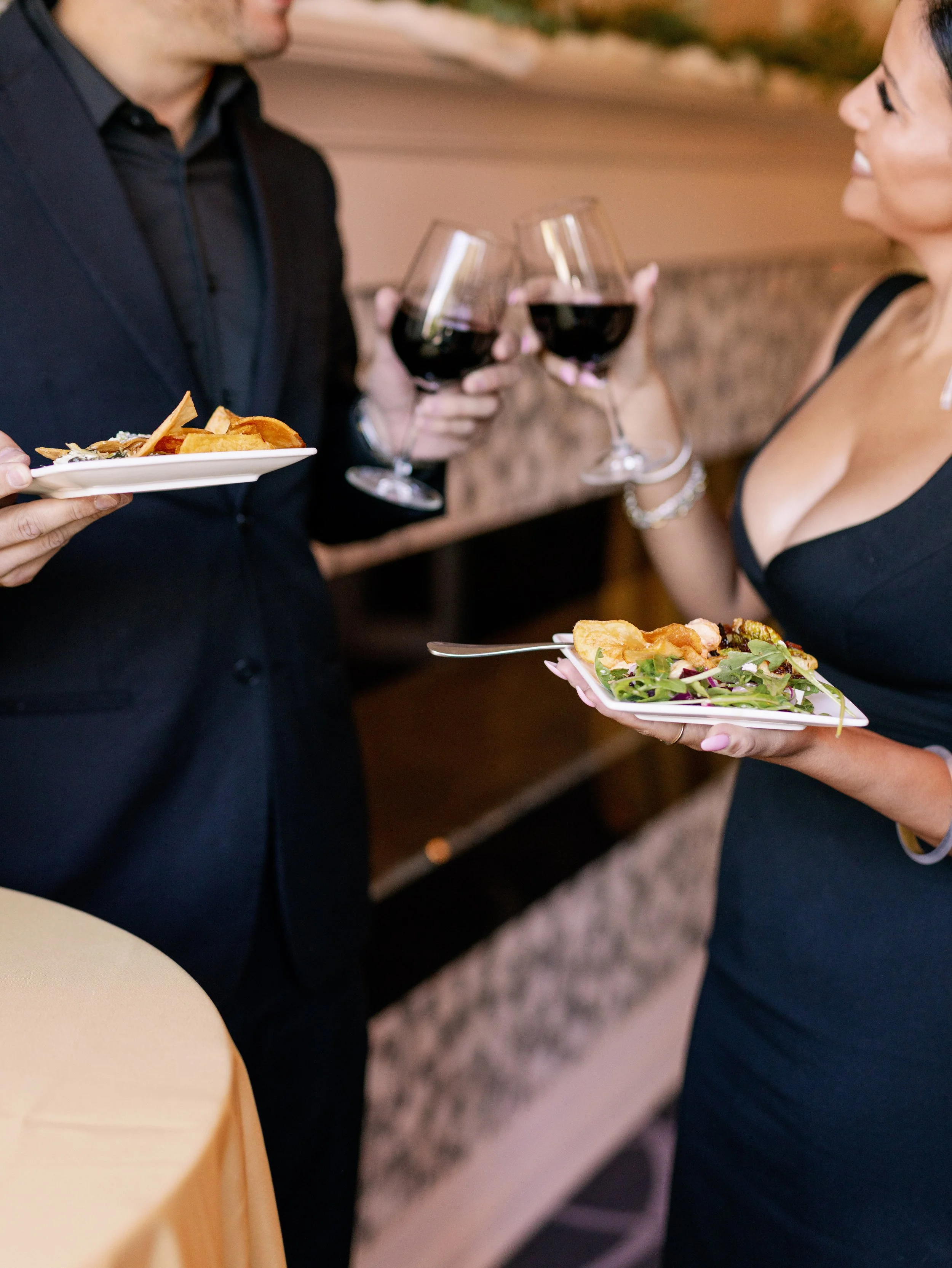 Two people are clinking glasses of red wine at a social gathering, with one person holding a plate of food, and the woman smiling and holding a plate of salad and other food items.