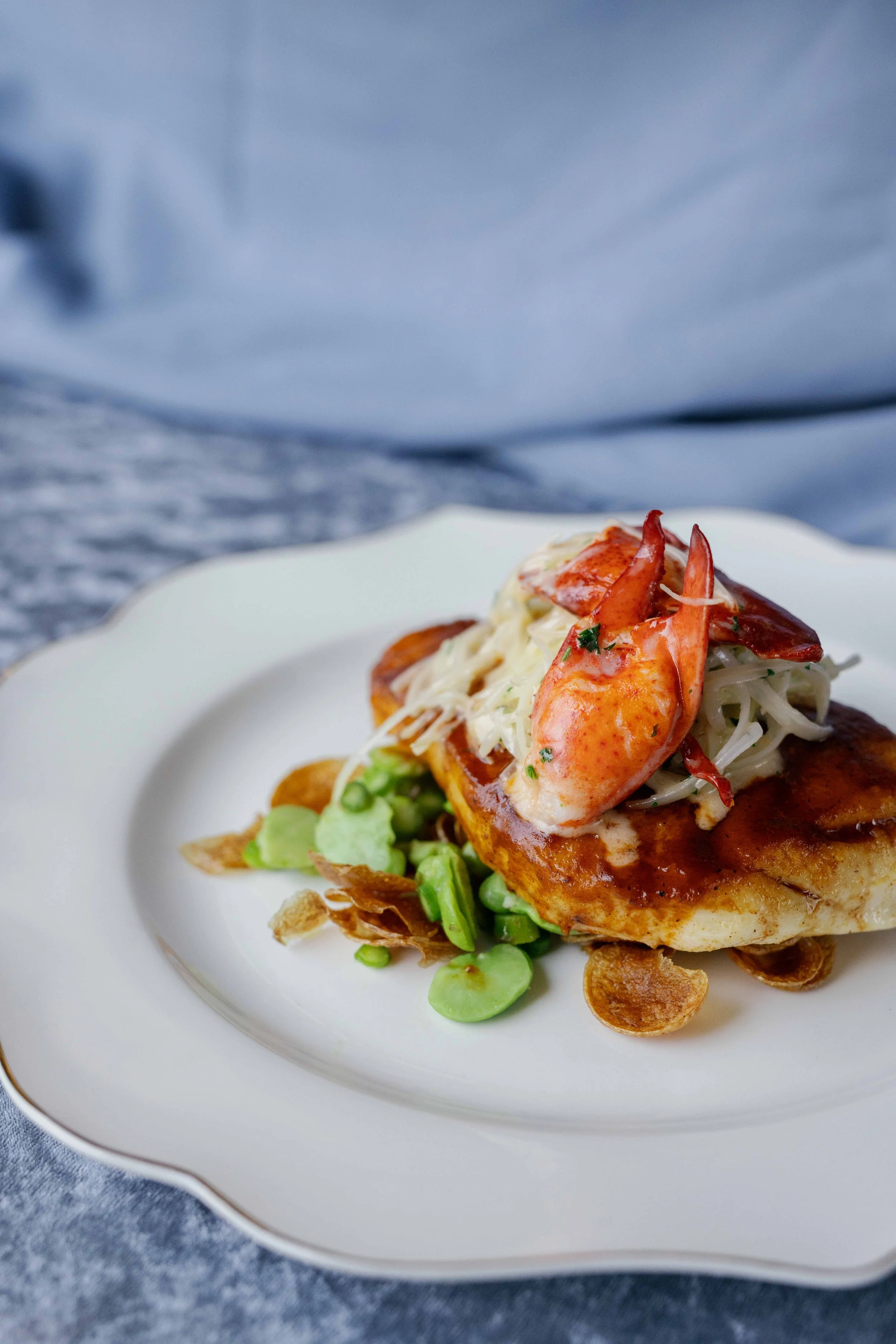 A plated dish featuring a piece of cooked chicken topped with lobster meat, shredded cheese, and sauce, garnished with green vegetables and crispy chips on a white plate.