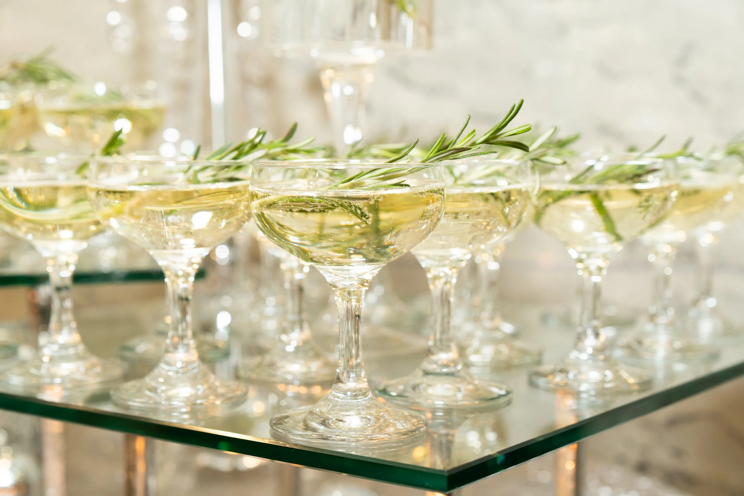 Clear cocktails with sprigs of fresh rosemary garnishing each drink, served in stemmed glasses, arranged on a mirrored tray.