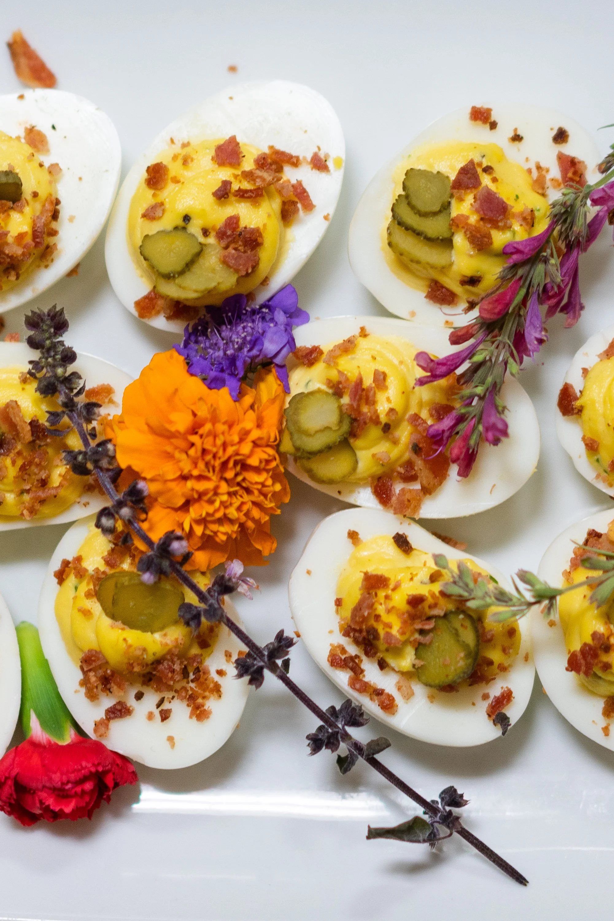 Deviled eggs garnished with bacon bits and pickle slices, arranged on a white surface with purple, orange, and red flowers.