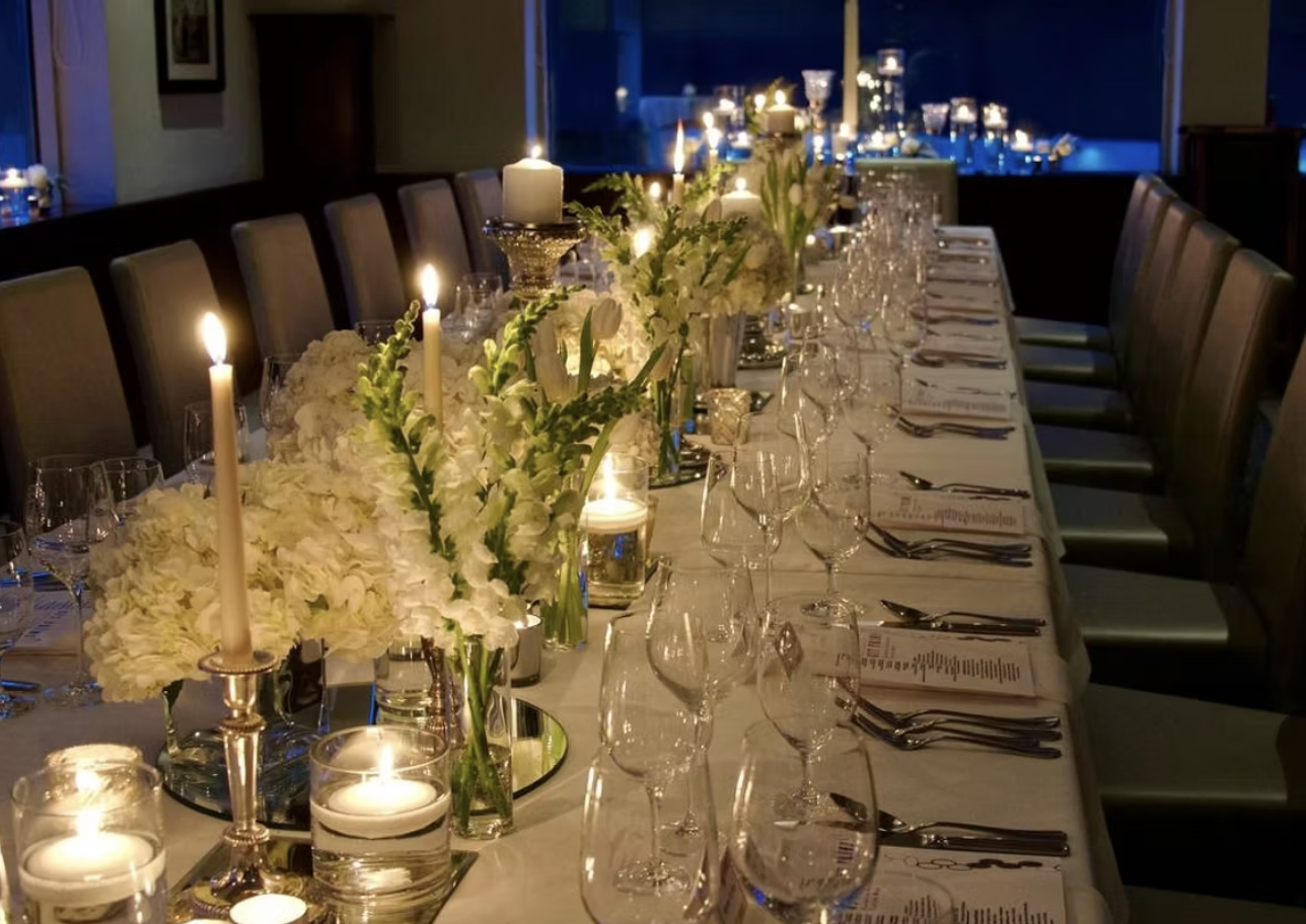 Elegant dining table set for a formal event with white floral centerpieces, candles, wine glasses, and utensils