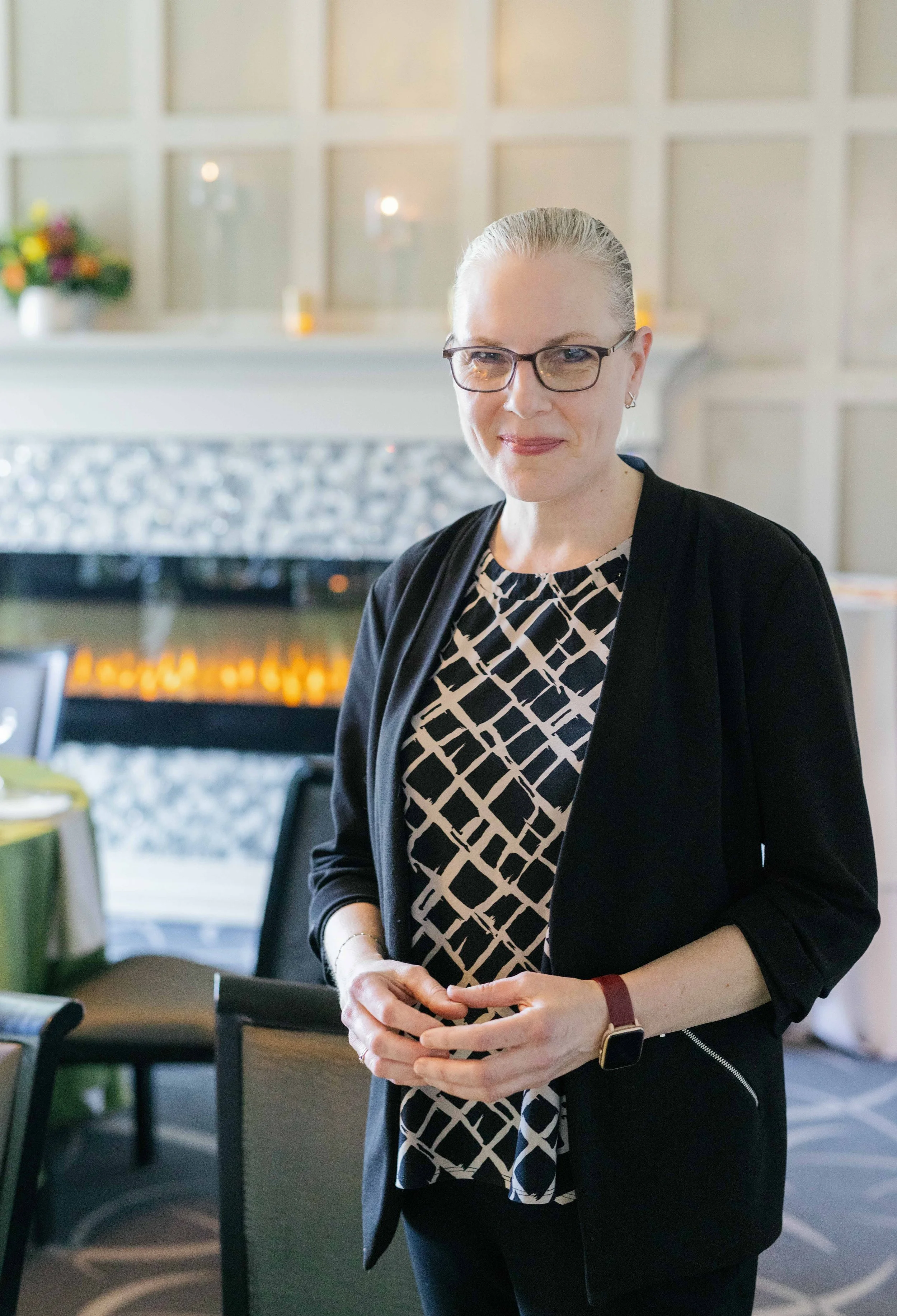 A woman with gray hair and glasses wearing a black blazer and patterned blouse, standing in a room with a fireplace and floral arrangement in the background.