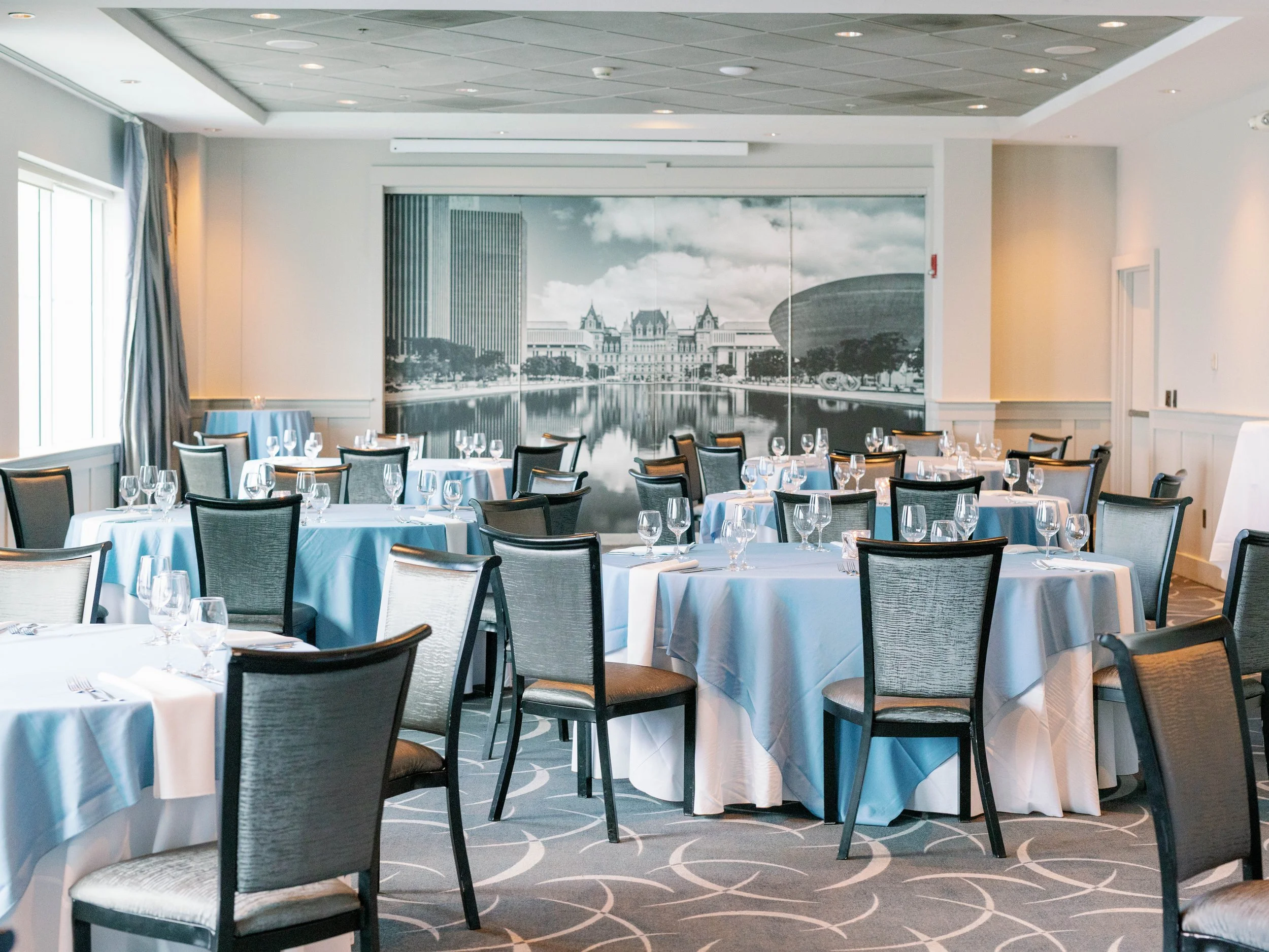 A banquet room set up with round tables covered in white tablecloths, each with wine glasses, plates, and silverware, and chairs arranged around them. The wall features a large black-and-white mural of a cityscape with water reflections and historic 