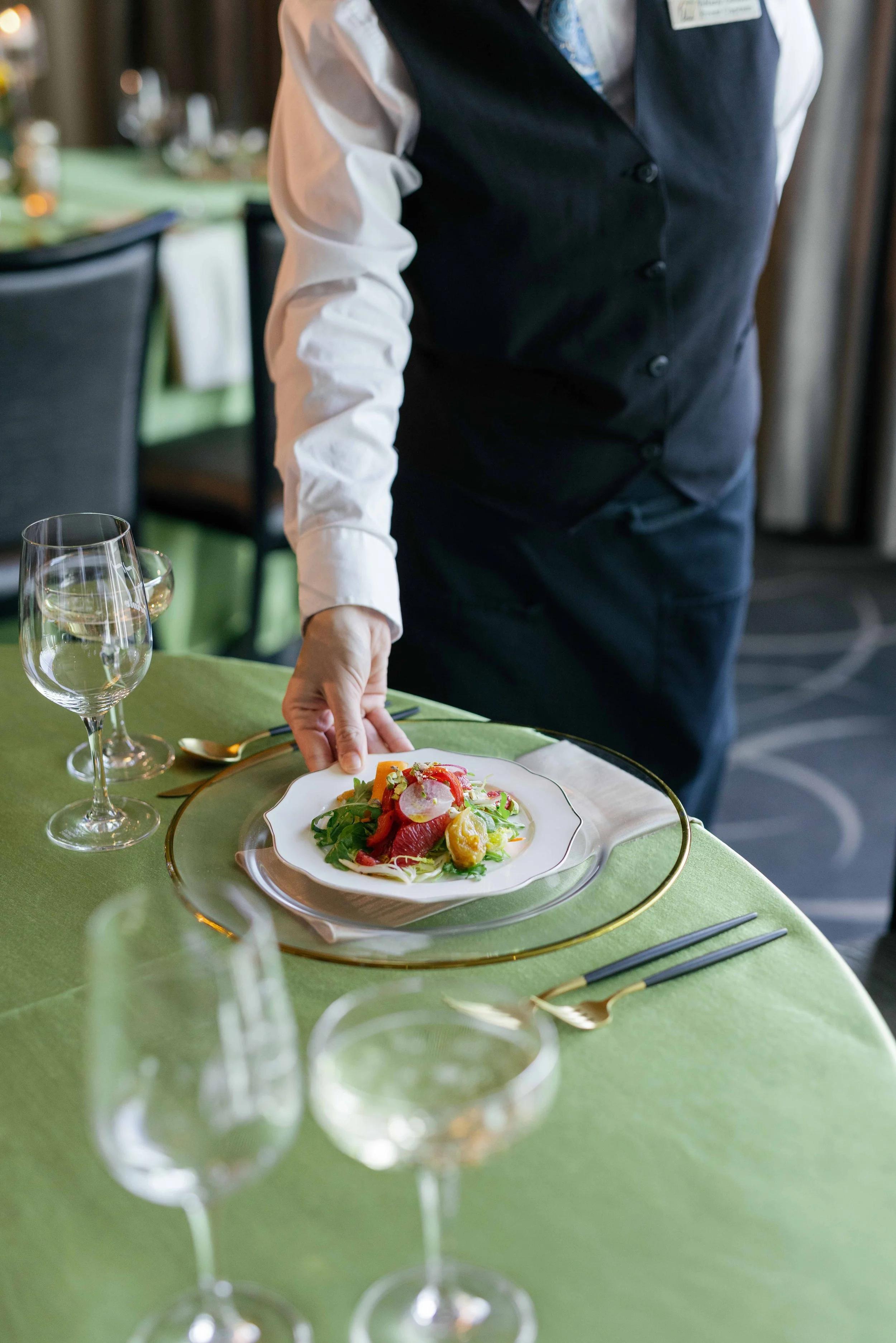 A waiter in a black vest and white shirt serving a plate of salad at a green-covered dining table in a restaurant.