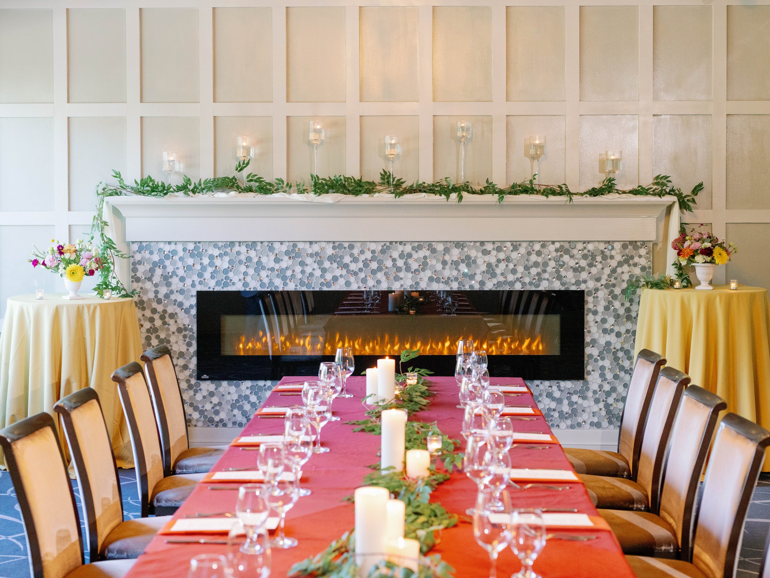 A decorated indoor dining area with a long table covered with a red tablecloth, set with glasses and napkins. The room features a white fireplace with candles and greenery on the mantel, and flower arrangements in vases on small tables on each side. 