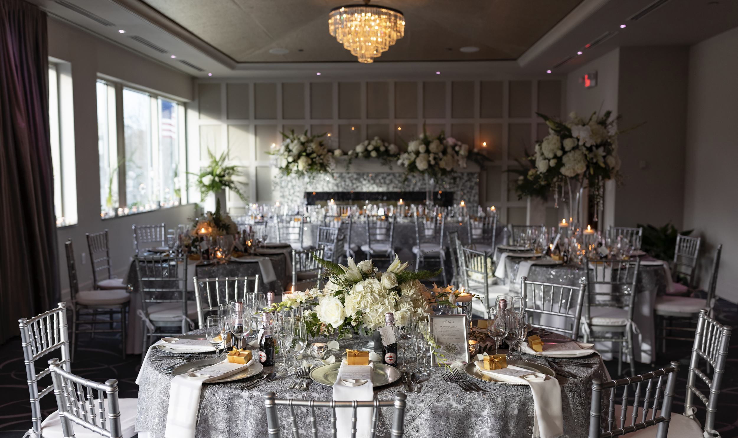 Elegant banquet hall decorated for a wedding reception with round tables, white floral centerpieces, candlelights, and silver chairs, with large windows and a chandelier on the ceiling.