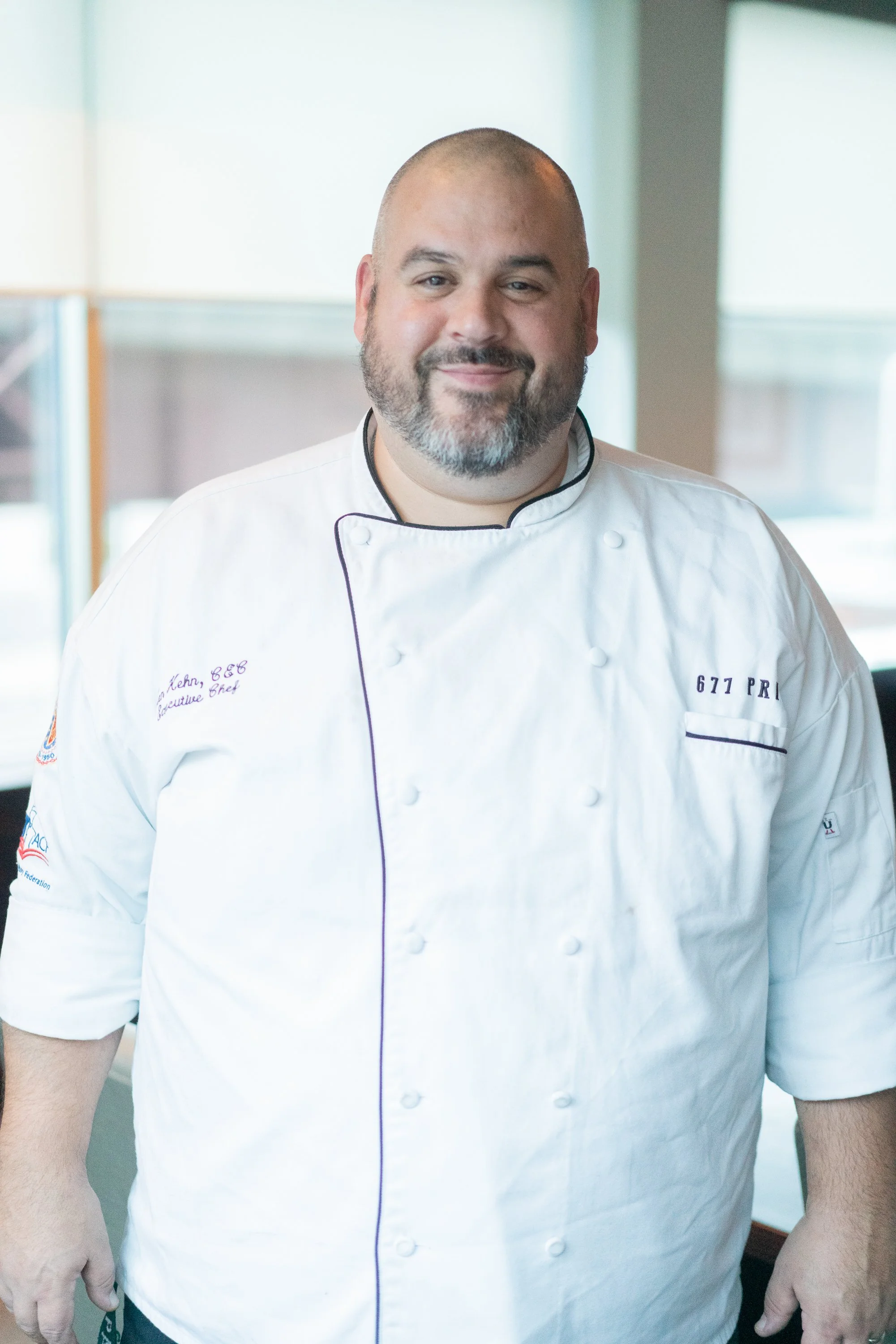 A male chef with a beard and shaved head wearing a white chef's jacket standing in a bright kitchen.