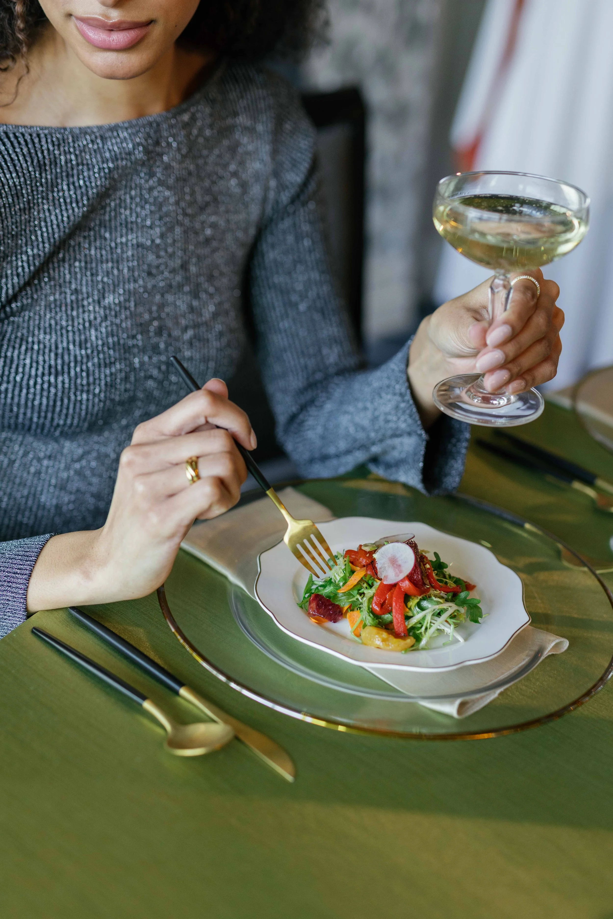 A woman in a sparkly gray dress is holding a glass of white wine in one hand and a fork in the other, sitting at a green table with a colorful salad on a white plate.