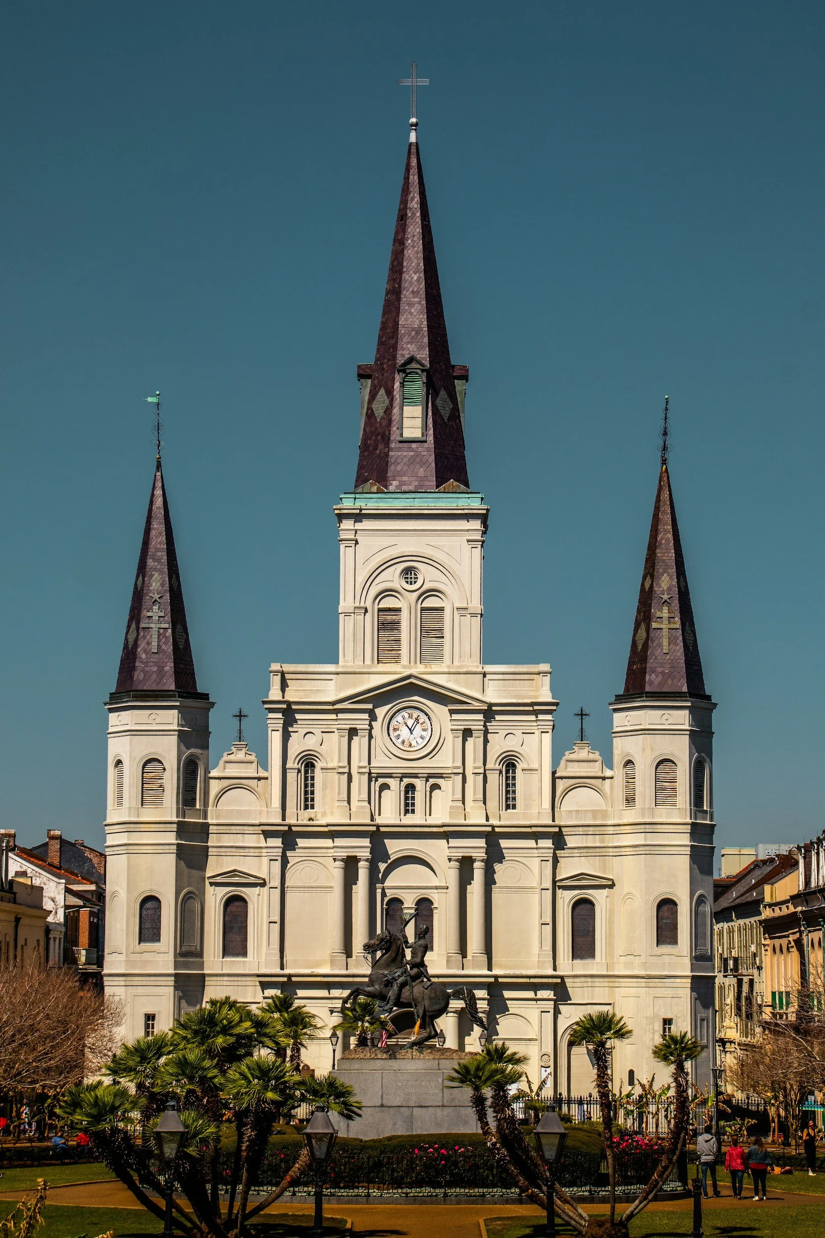 Statue of a man on horseback in front of a large white church with three tall, pointed steeples, set in a town square with palm trees and people walking around.