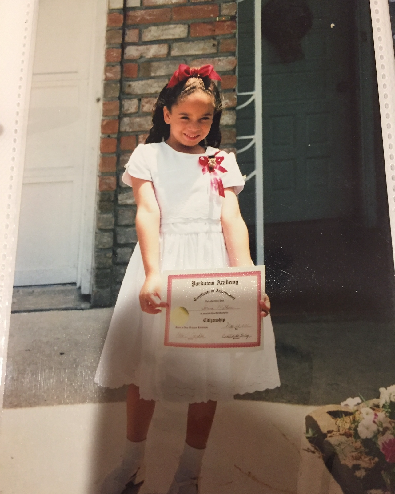 A young girl in a white dress with a red bow in her hair, holding a certificate of achievement outdoors in front of a brick wall. She is smiling.