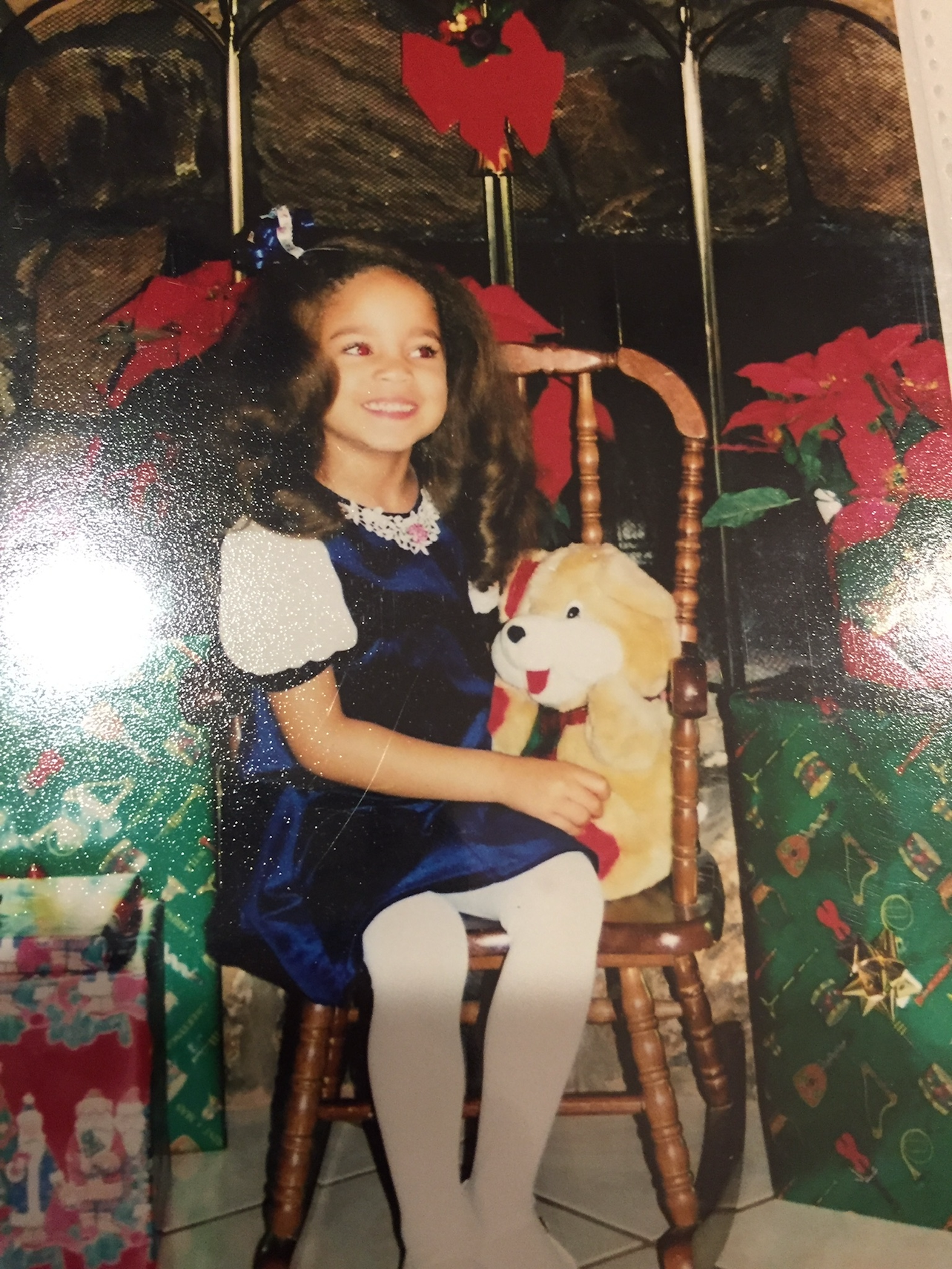 A young girl sitting on a wooden chair, holding a plush dog toy, surrounded by Christmas presents and decorations, including poinsettias and gift bags.