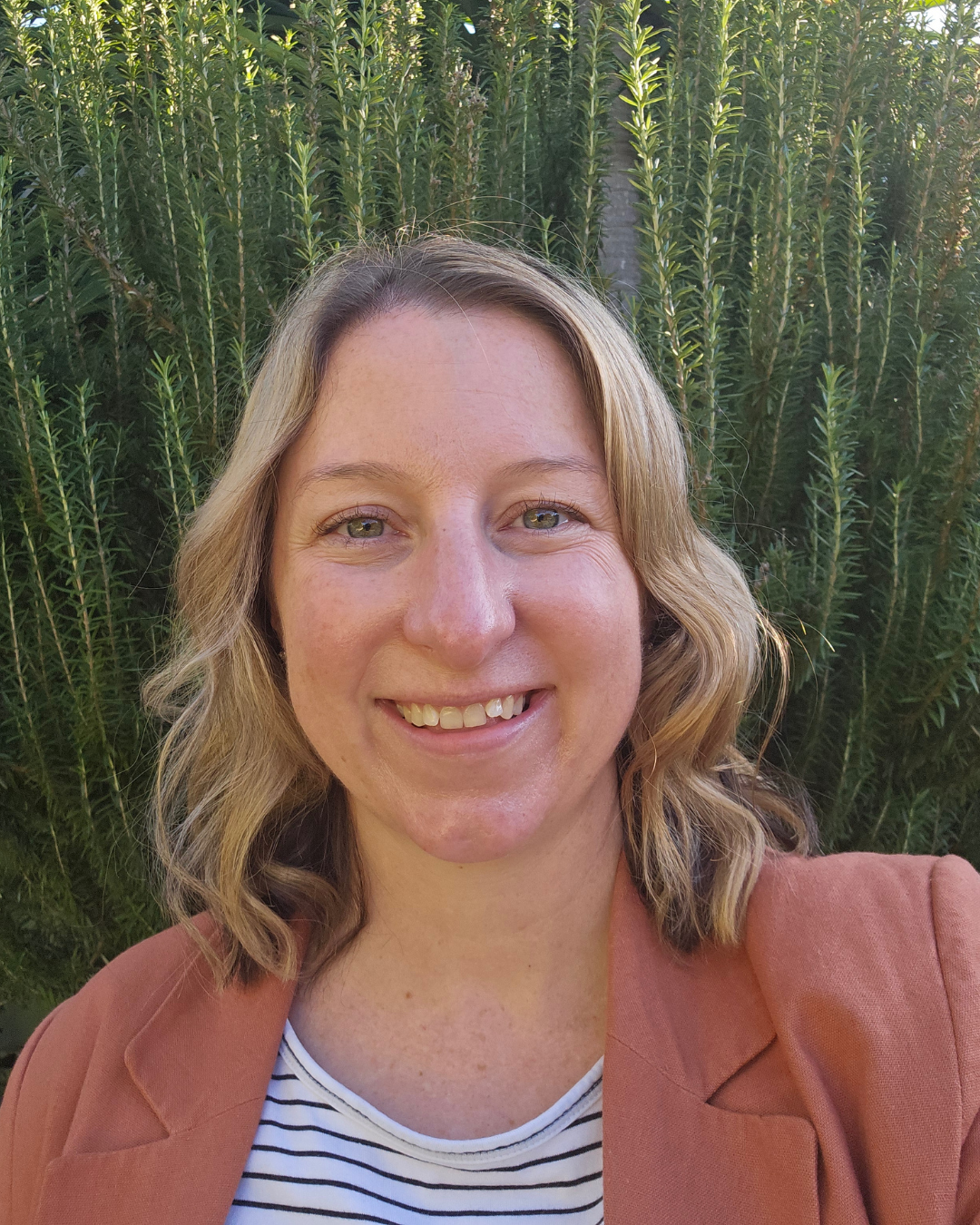 Close-up of a smiling woman with shoulder-length wavy blonde hair, wearing a pink blazer and white striped top, standing in front of green foliage.