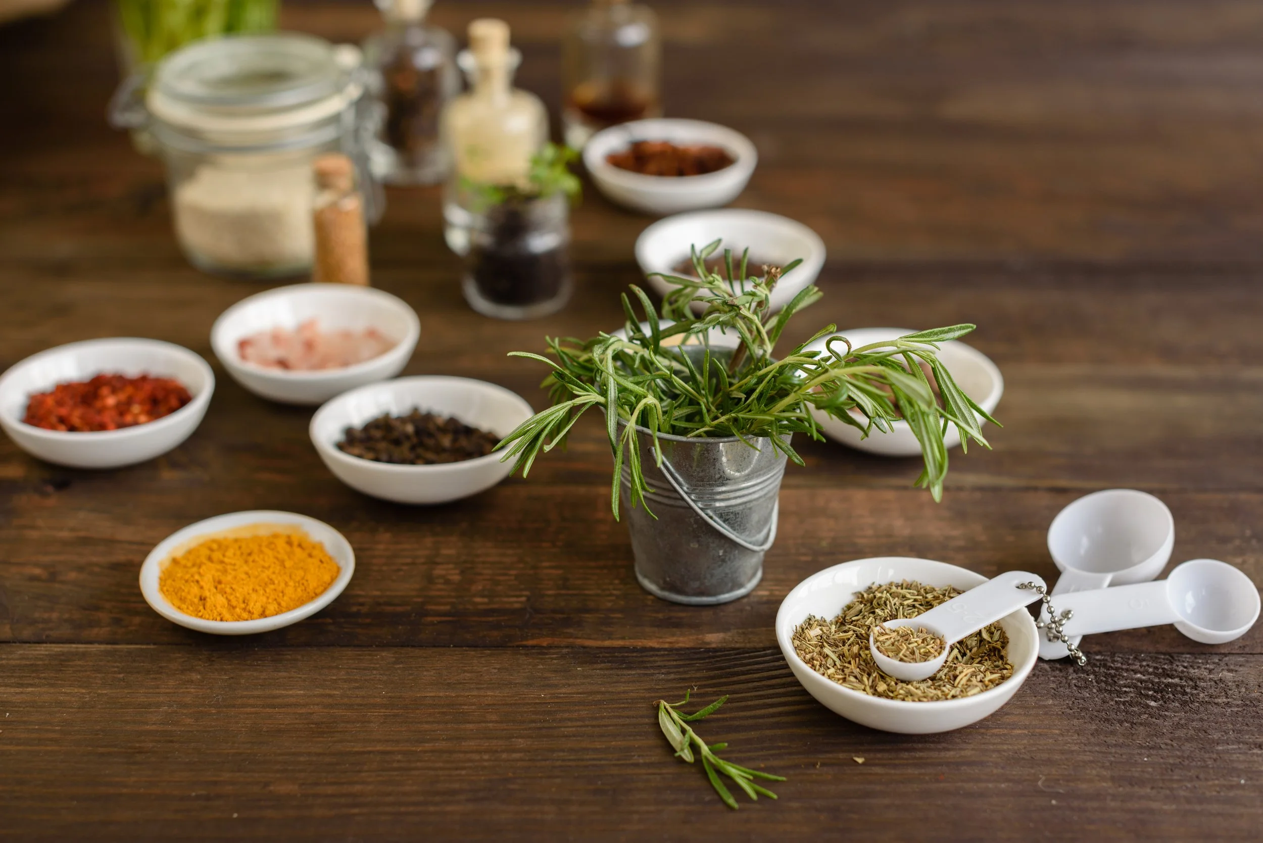 A small potted rosemary plant on a wooden table surrounded by bowls of various herbs and spices, including turmeric, dried herbs, chili flakes, and other seasonings.