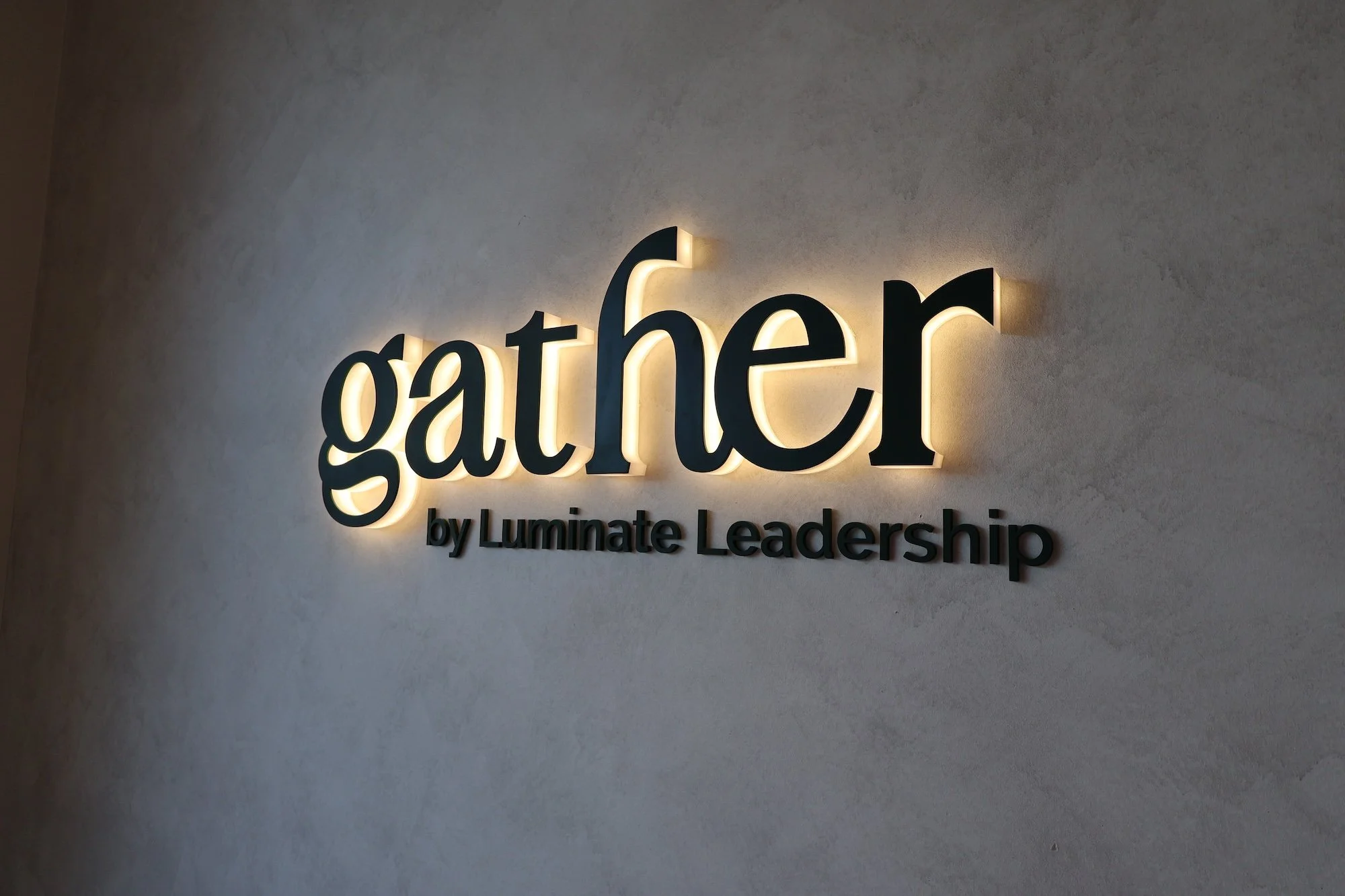 Backlit sign that reads 'gather' with smaller text below saying 'by Luminate Leadership,' mounted on a plain textured wall.