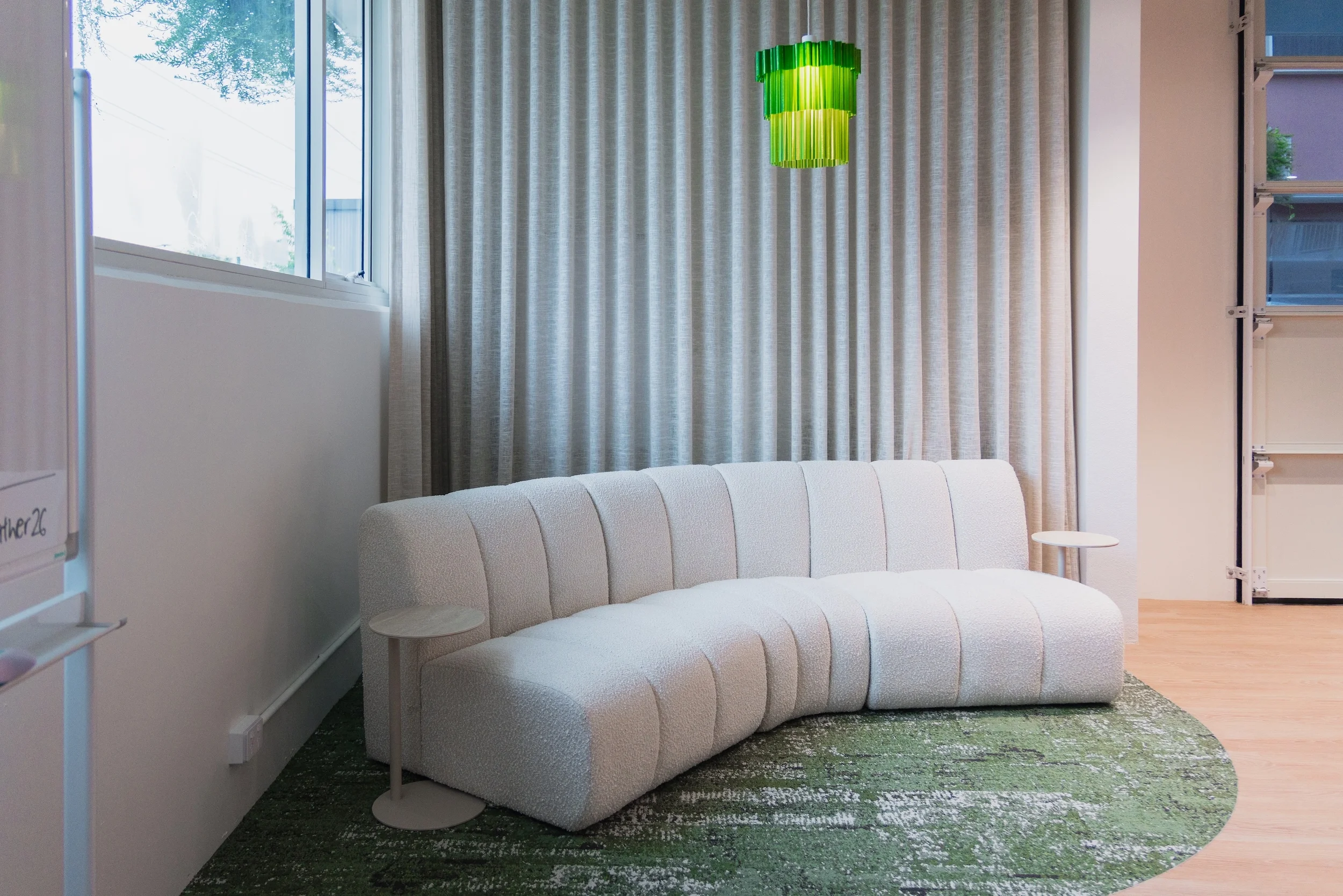 A modern living room with a white curved sofa, a small round side table, a large window with sheer curtains, and a bright green hanging light fixture.