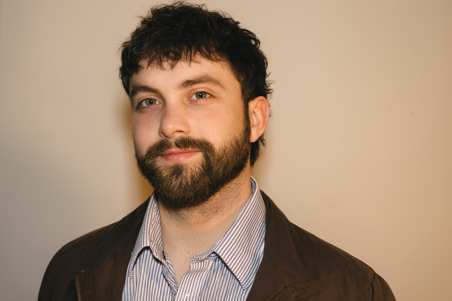 A man with dark, slightly curly hair, blue eyes, and a beard, dressed in a striped shirt and brown jacket, standing against a plain, light-colored wall.