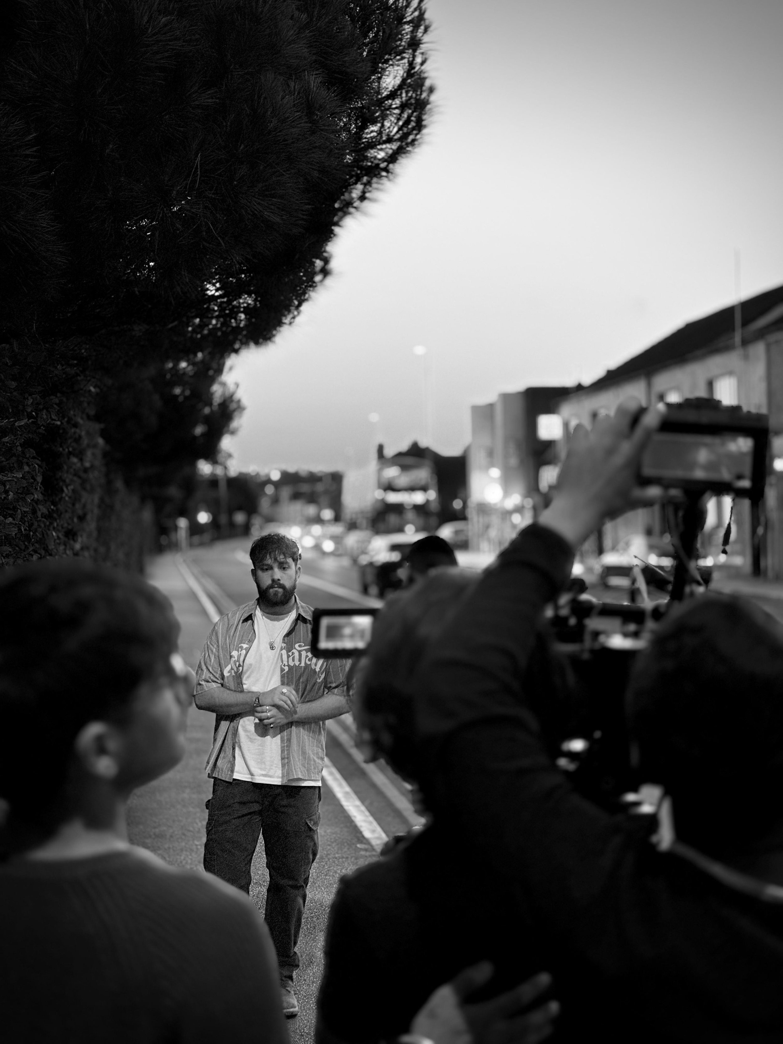 A man is being filmed on a city street during twilight, with a small crew around him capturing the scene.