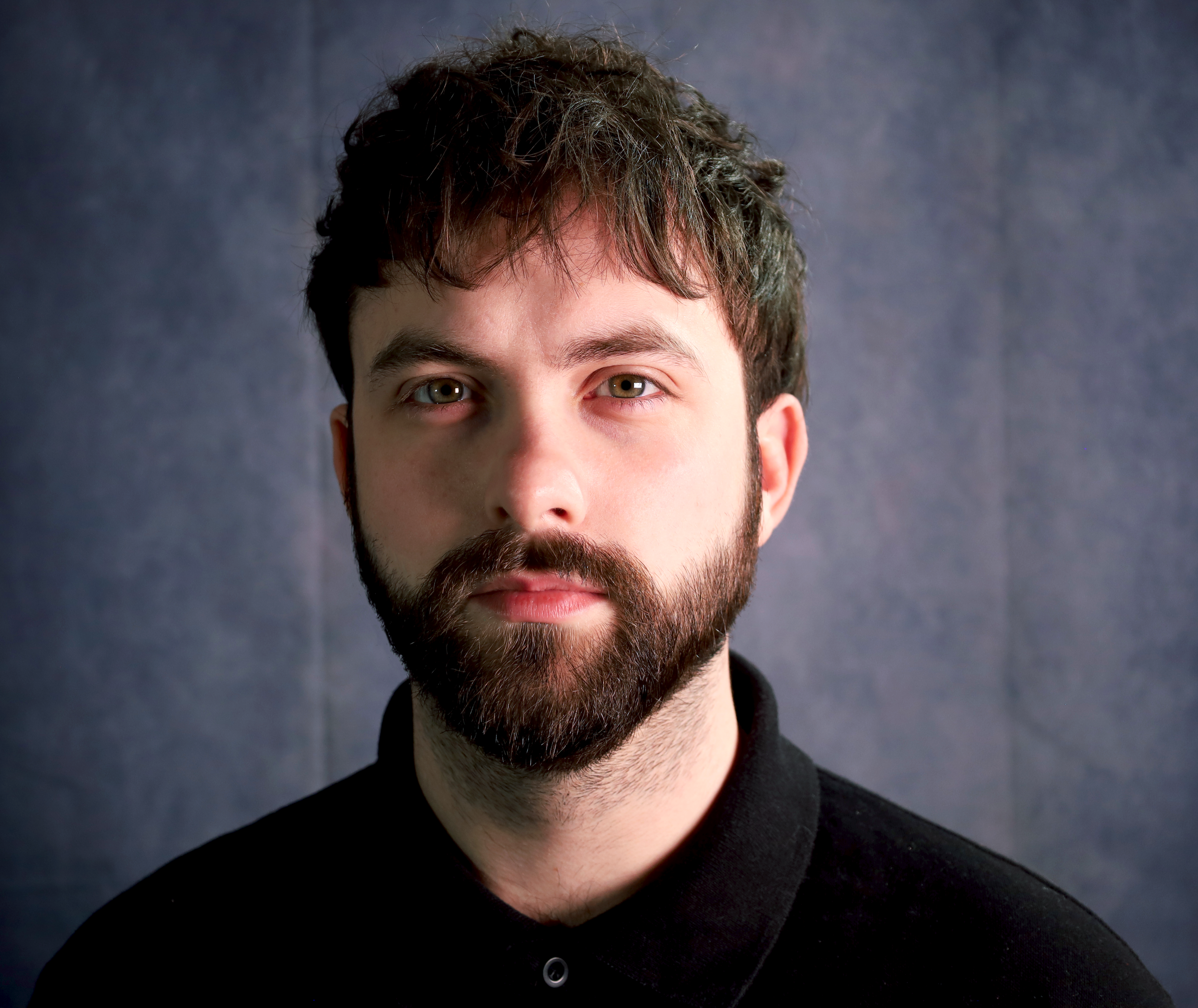 Close-up portrait of a man with brown hair, beard, and light-colored eyes, wearing a black shirt against a dark background.