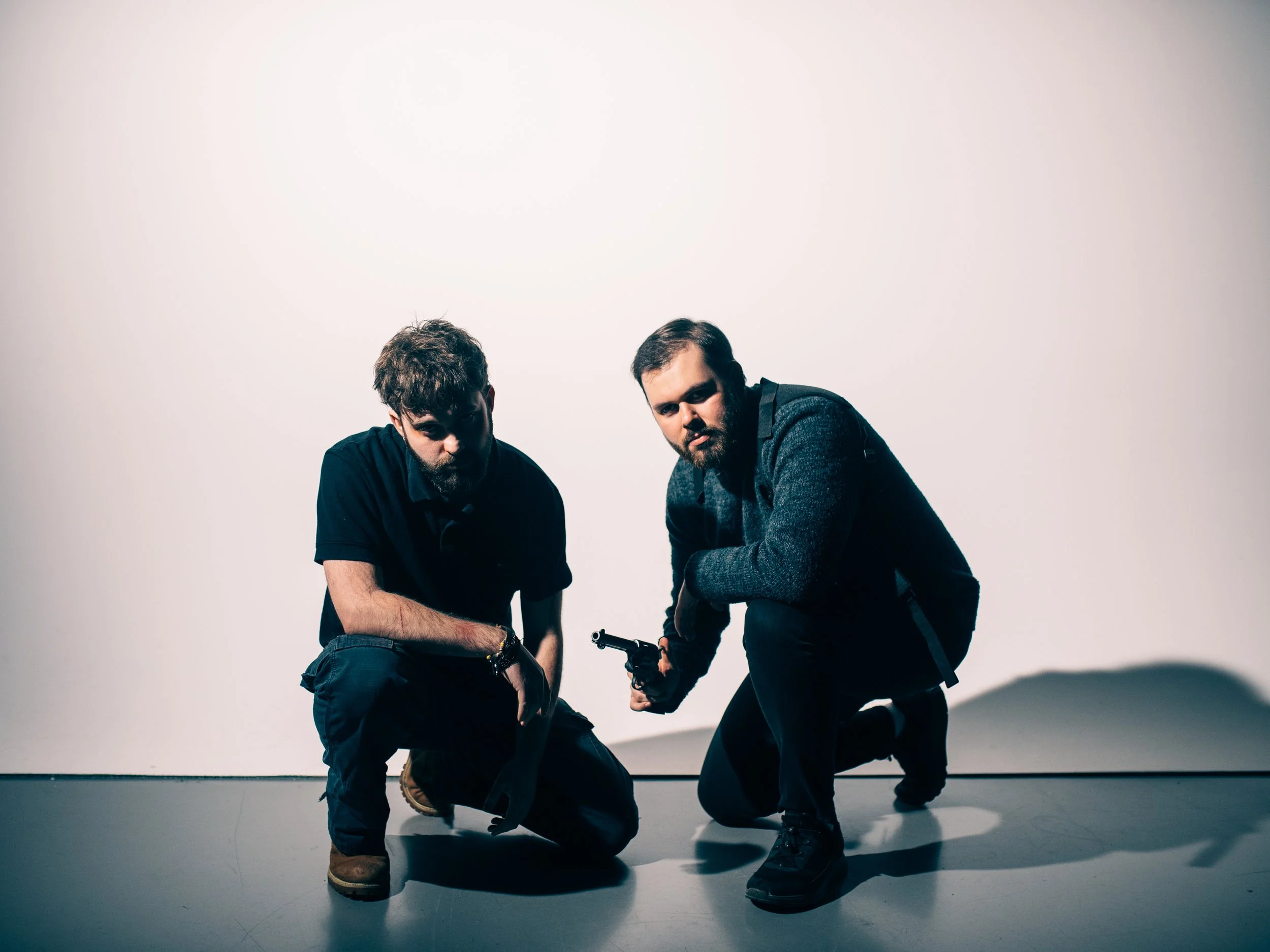 Two men kneeling on the floor, one holding a handgun, in a studio with plain white background.