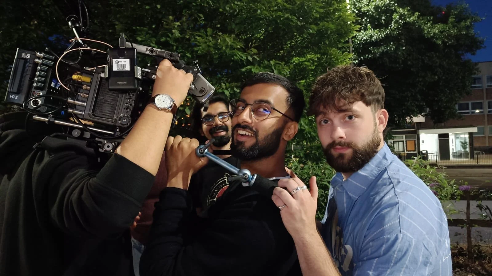 Three men, with one holding a professional film camera, pose for the photo outdoors at night with greenery and a building in the background.