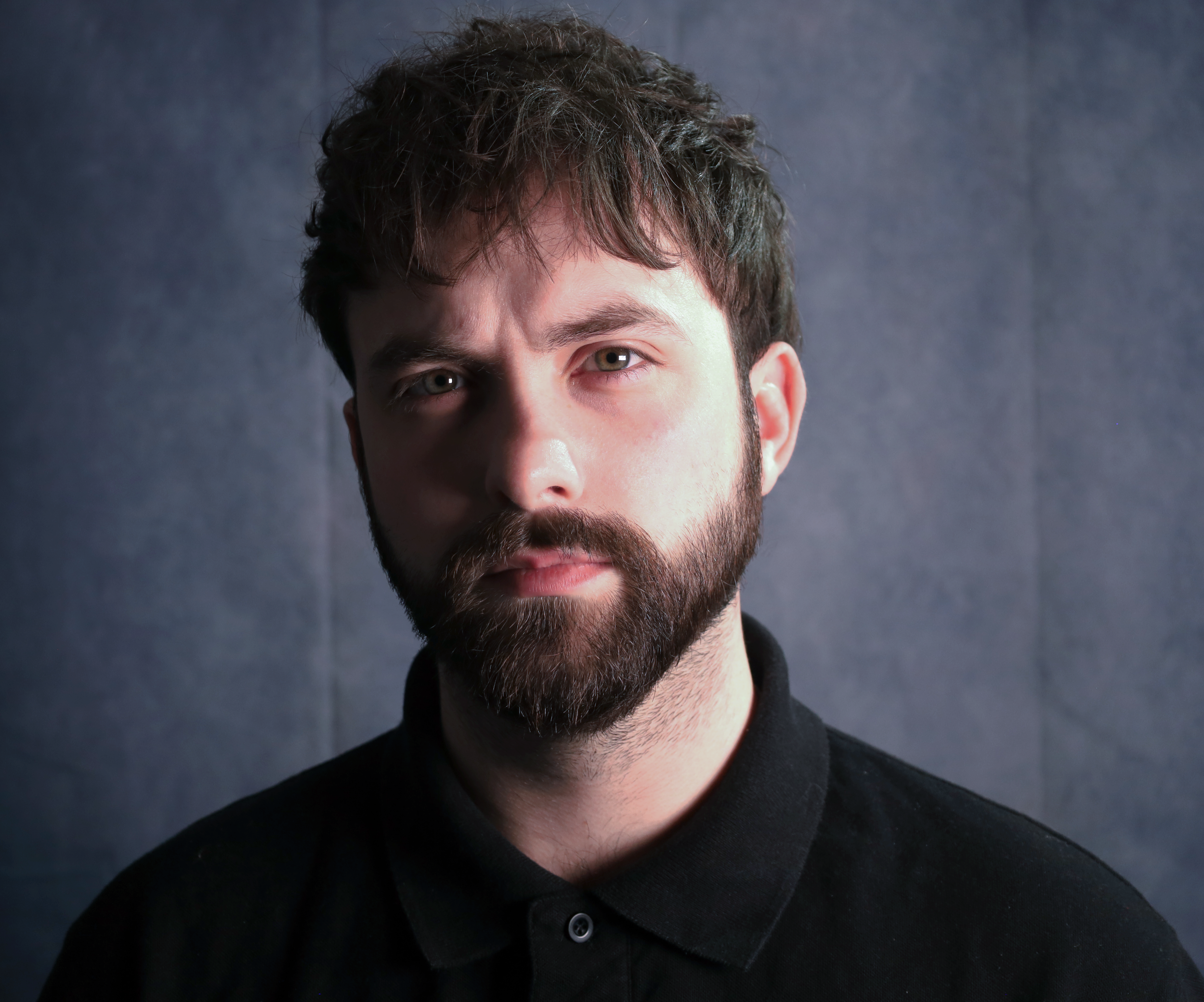 A portrait of a young man with dark hair and a beard, wearing a black shirt, against a dark gray background.