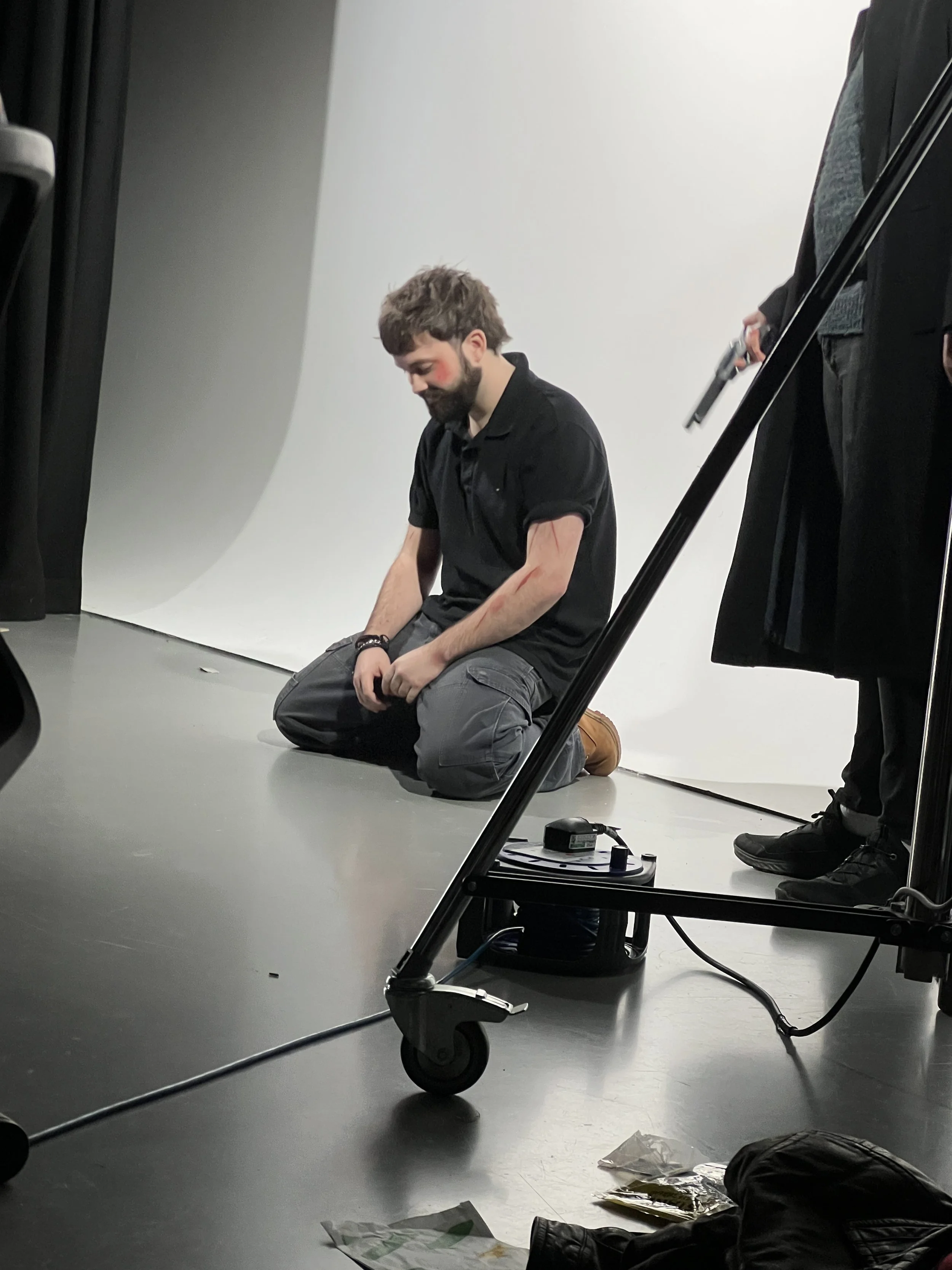 A man kneeling on the floor with fake injuries and blood marks on his arms and face, participating in a photoshoot or scene in front of a white backdrop, with photography equipment nearby.