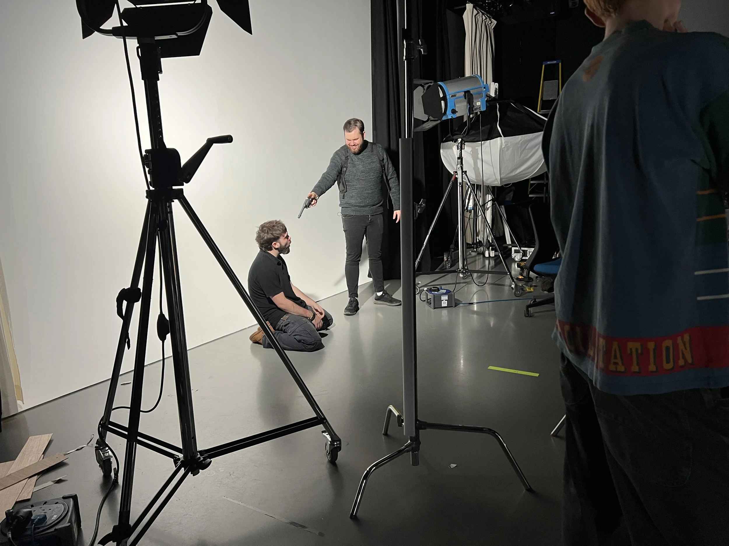Behind-the-scenes photo of a video shoot in a studio, with a man kneeling and a man standing, pointing a gun prop at him, surrounded by lighting and camera equipment.
