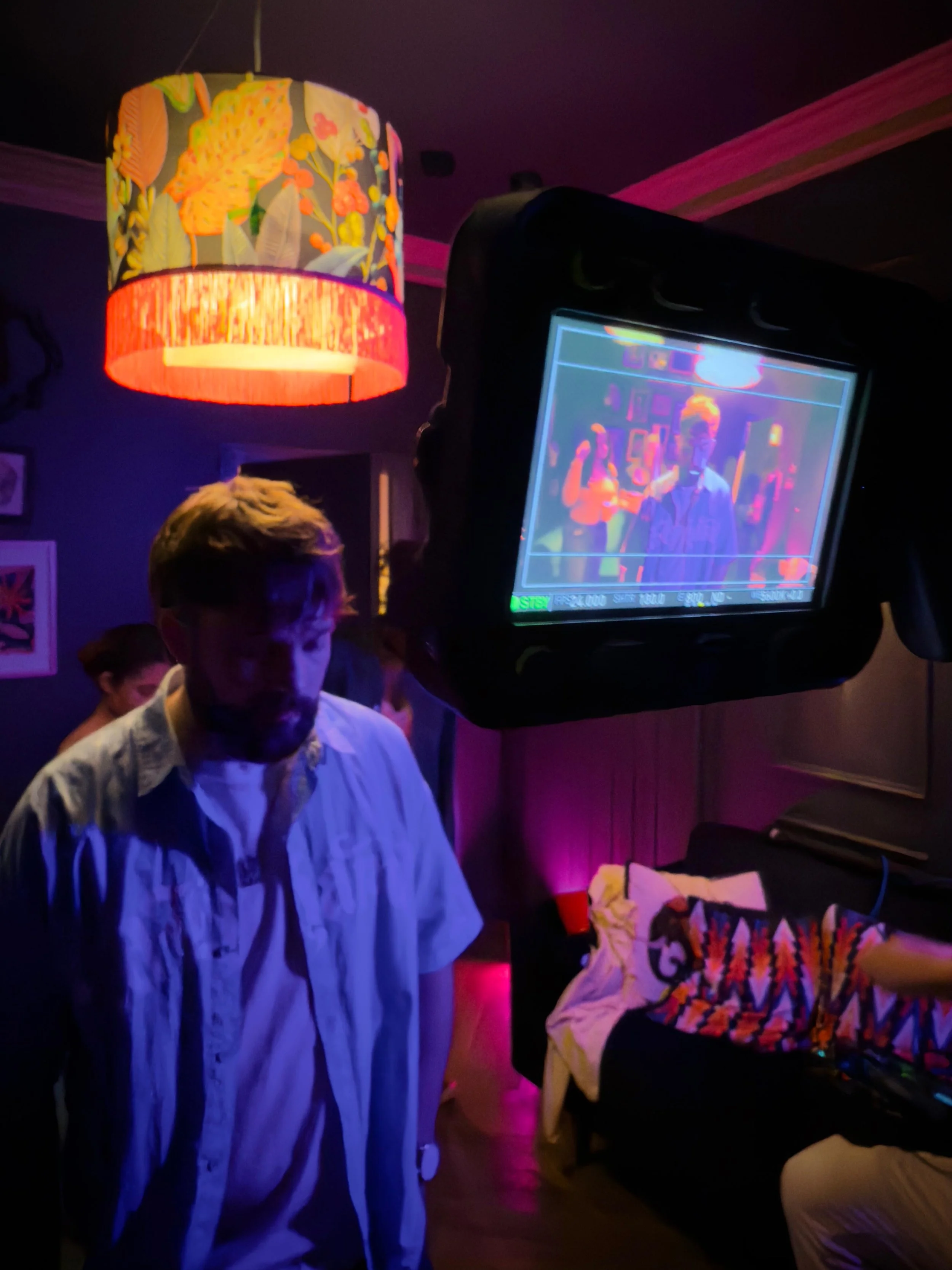 A person with light brown hair and a beard stands in a dimly lit room with purple and pink lighting. A large, colorful lampshade with floral patterns hangs from the ceiling. On the right, a monitor displays a colorful image of the person in front of 