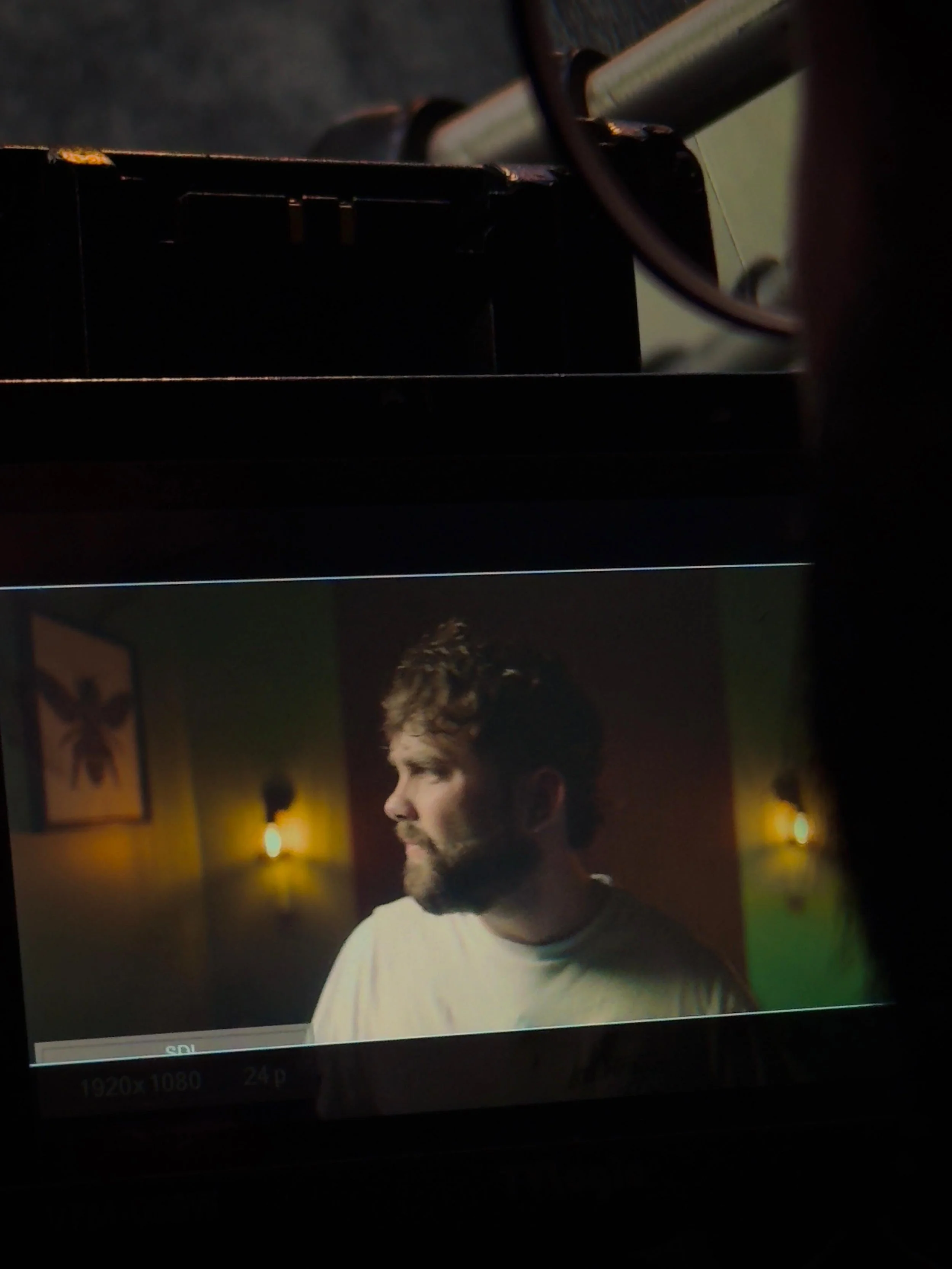 A man with brown hair and a beard, wearing a white shirt, is looking to his left inside a room with dim lighting.