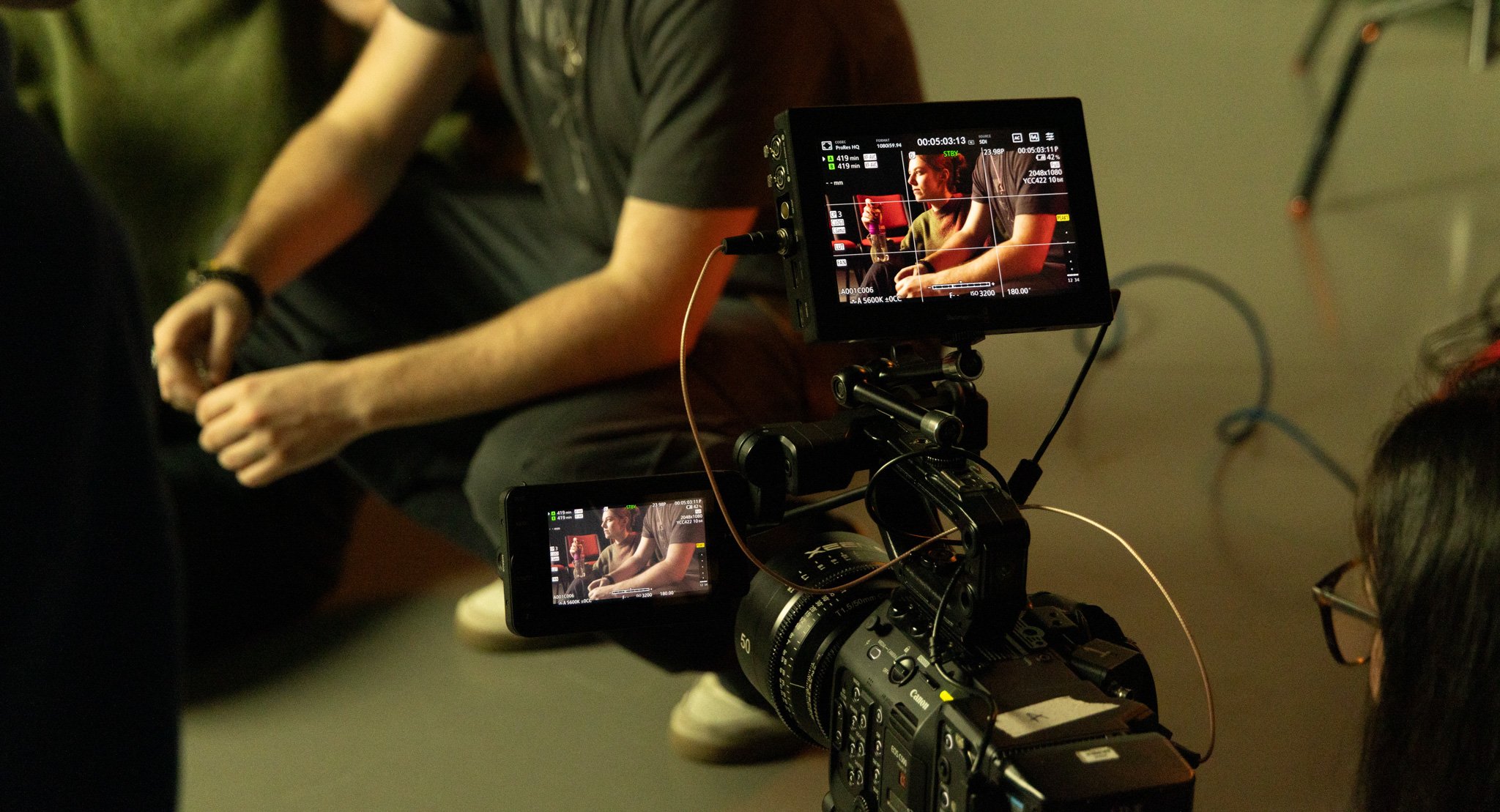 Camera filming a person sitting on a chair in a dimly lit indoor setting.
