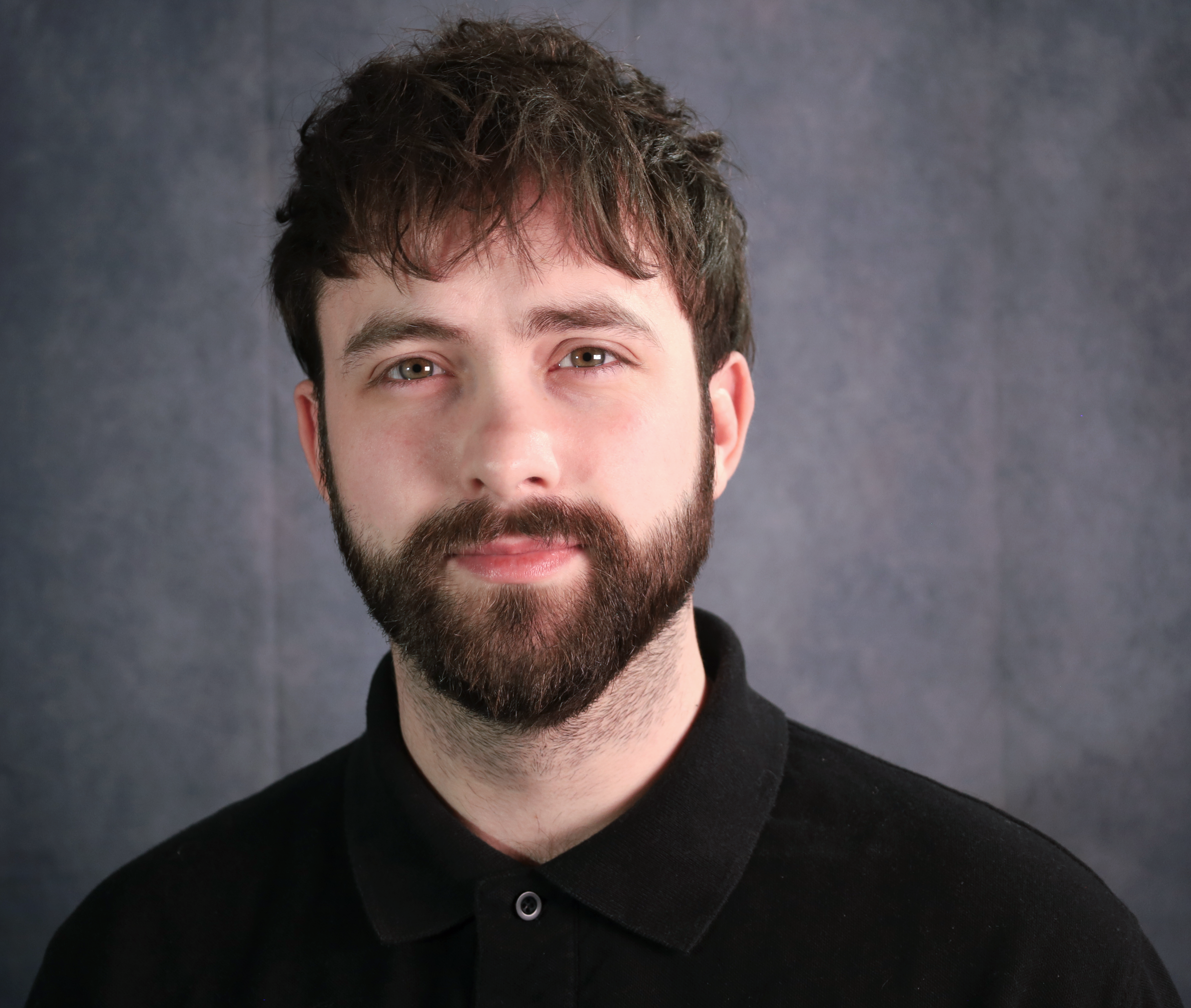 Close-up portrait of a young man with brown hair, beard, and blue eyes wearing a black collared shirt against a dark grey background.