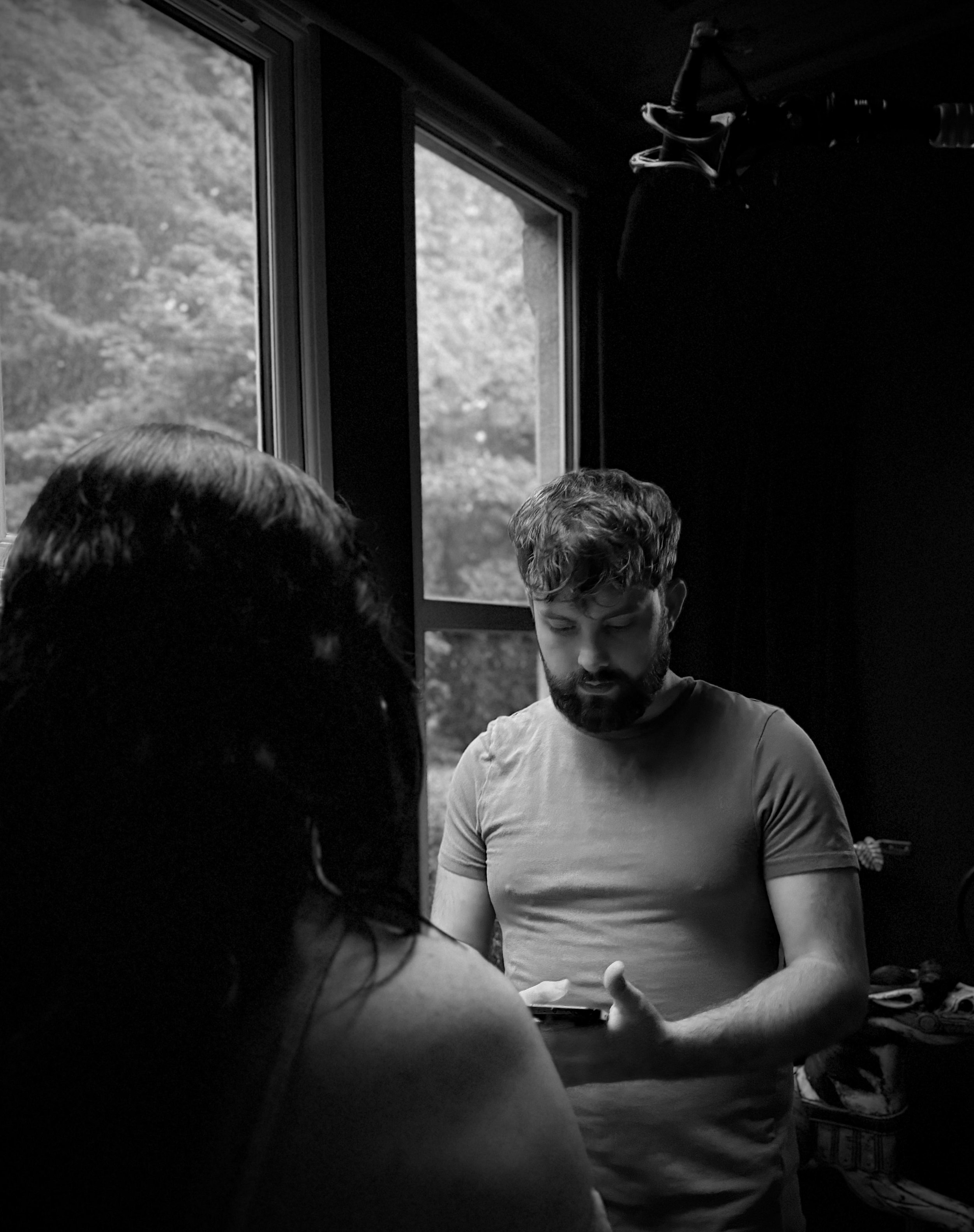 A man with a beard looking at his phone indoors, with a woman with glasses in front of him. There is a window with trees outside and a dark background.