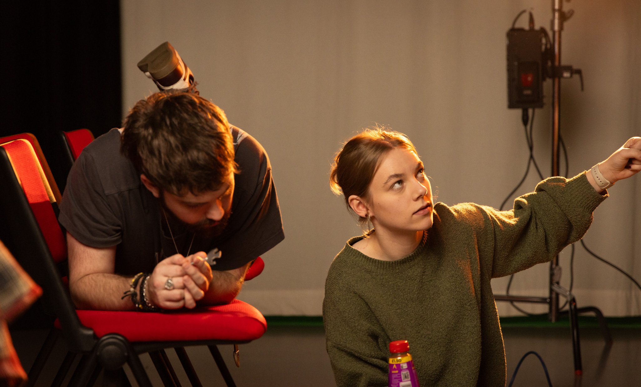 A young woman with brown hair, wearing a green sweater, is pointing or directing at something off-camera. Behind her, a young man with brown hair and wearing a dark gray shirt is leaning on a red and black chair, looking downward. There is a water bo