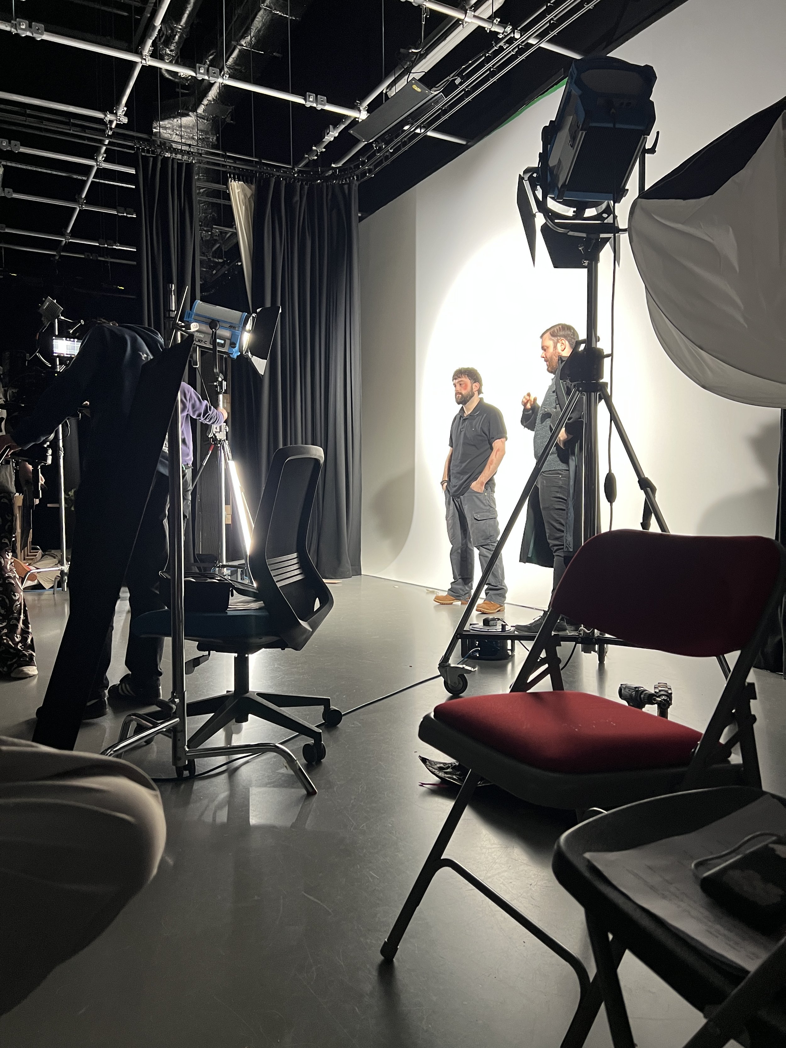 Two men are standing in front of a white backdrop in a studio, talking and gesturing. The studio has various filming equipment, lights, and chairs, with black curtains and ceiling piping visible.