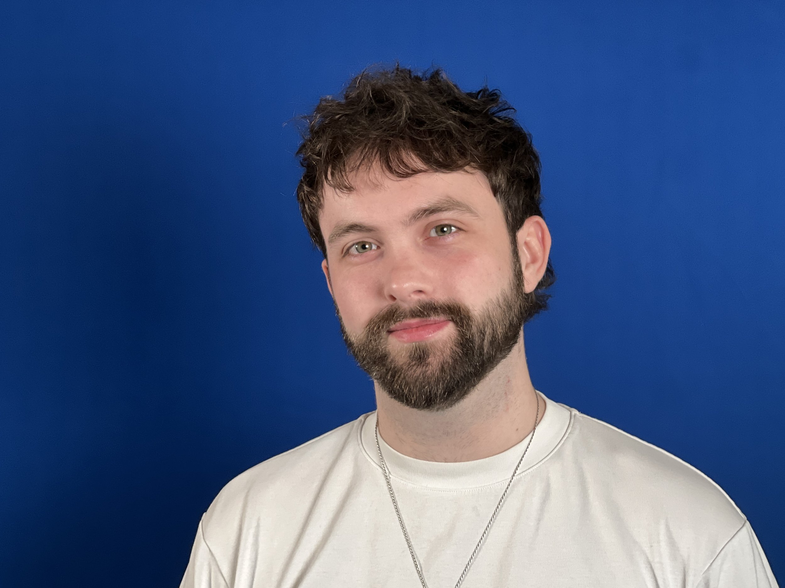 A young man with curly brown hair and a beard, wearing a white t-shirt and a silver chain necklace, poses against a solid blue background.