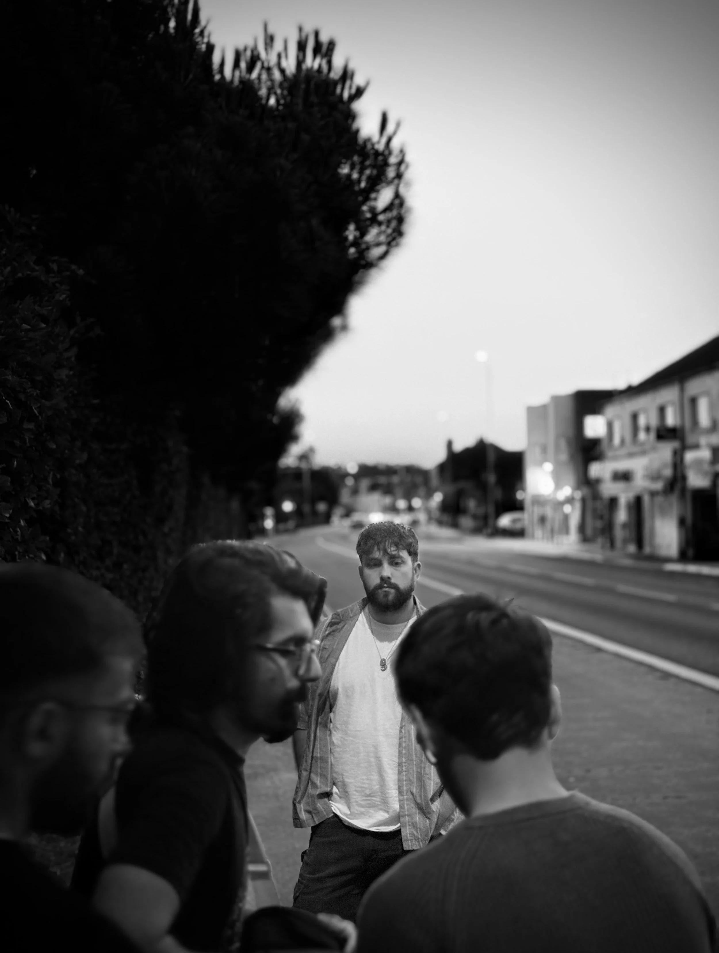 A black-and-white photo of four people gathered outdoors on a sidewalk, with one man standing apart from the group looking at them. The background shows a blurred street with buildings and streetlights.