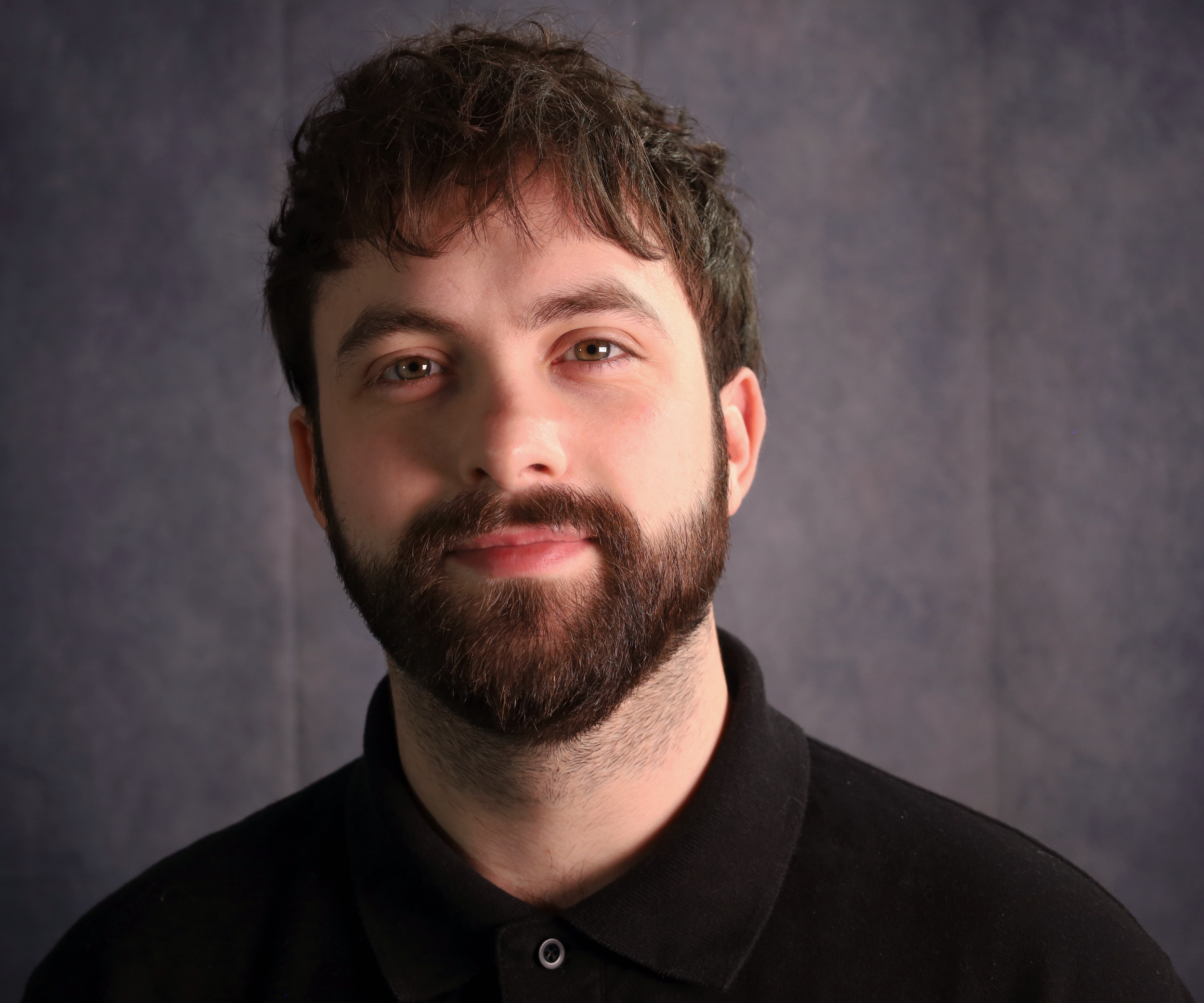 A portrait of a man with brown hair and a beard, wearing a black shirt, against a gray background.